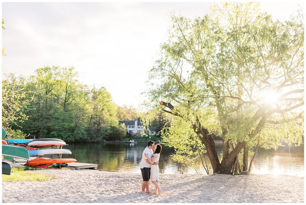 Couple shares a kiss on the private beach in their lakeside community during their Wayne New Jersey Engagement Photos. The sunlight is glistening through the trees.