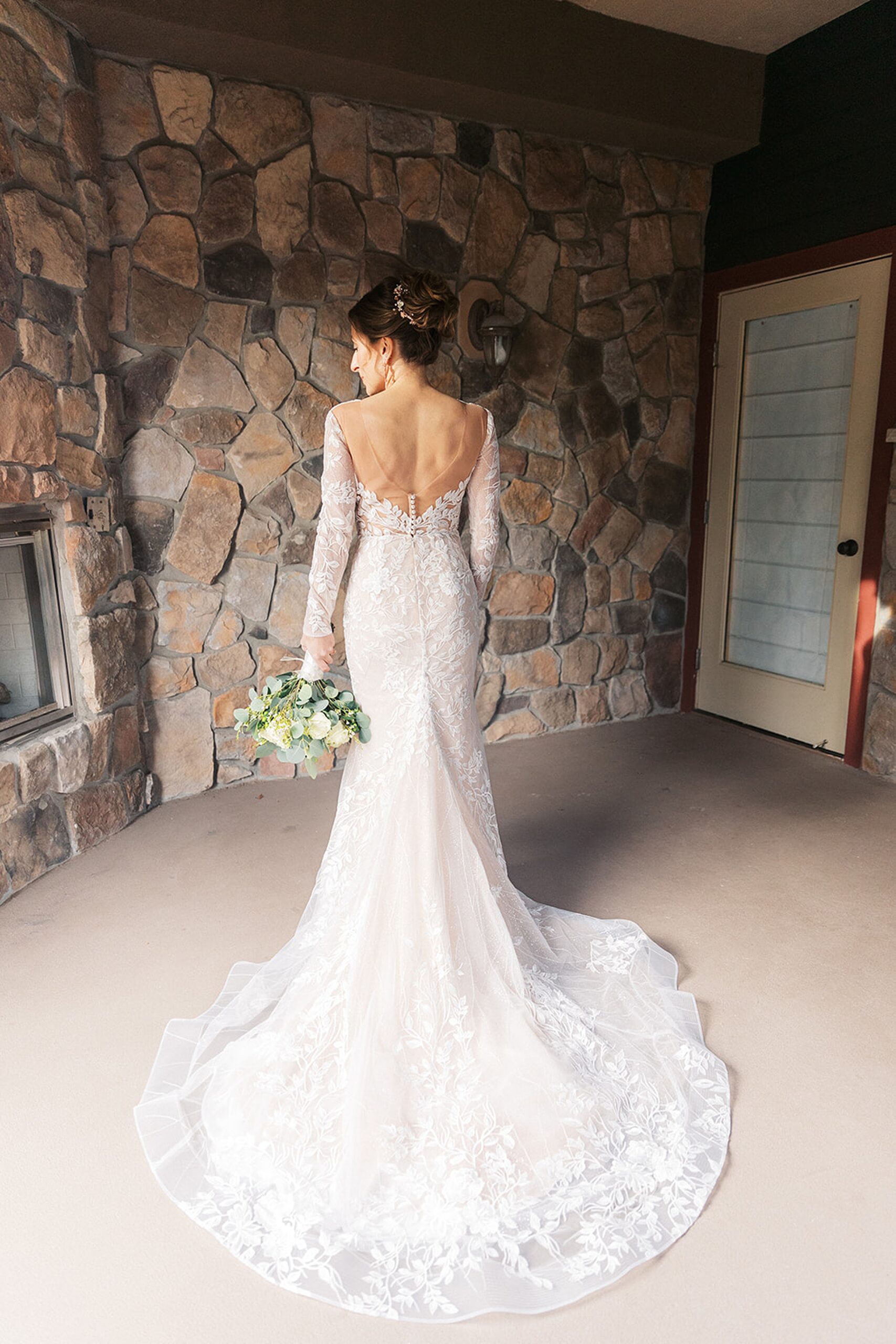 A bride stands by a stone fireplace in her white lace dress with bouquet held down by her side