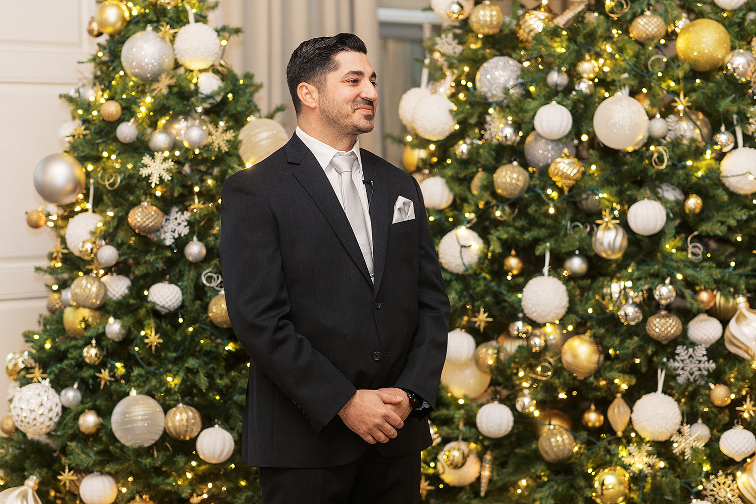 A groom smiles in front of christmas trees with gold and white ornaments while wearing a black suit and silver tie