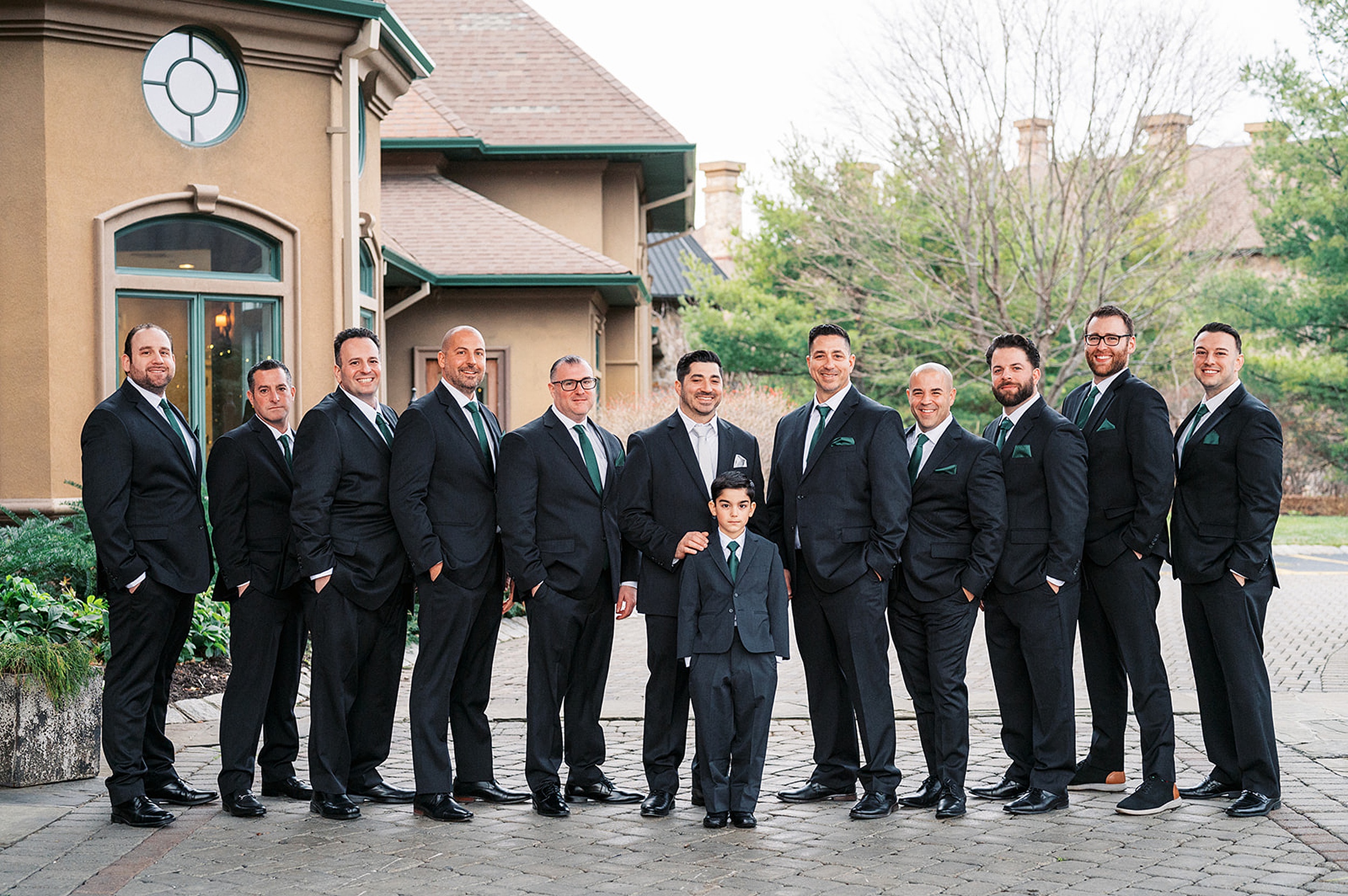 A groom stands on a stone patio in a black suit and white tie with his large party of groomsmen at a Crystal Springs Wedding