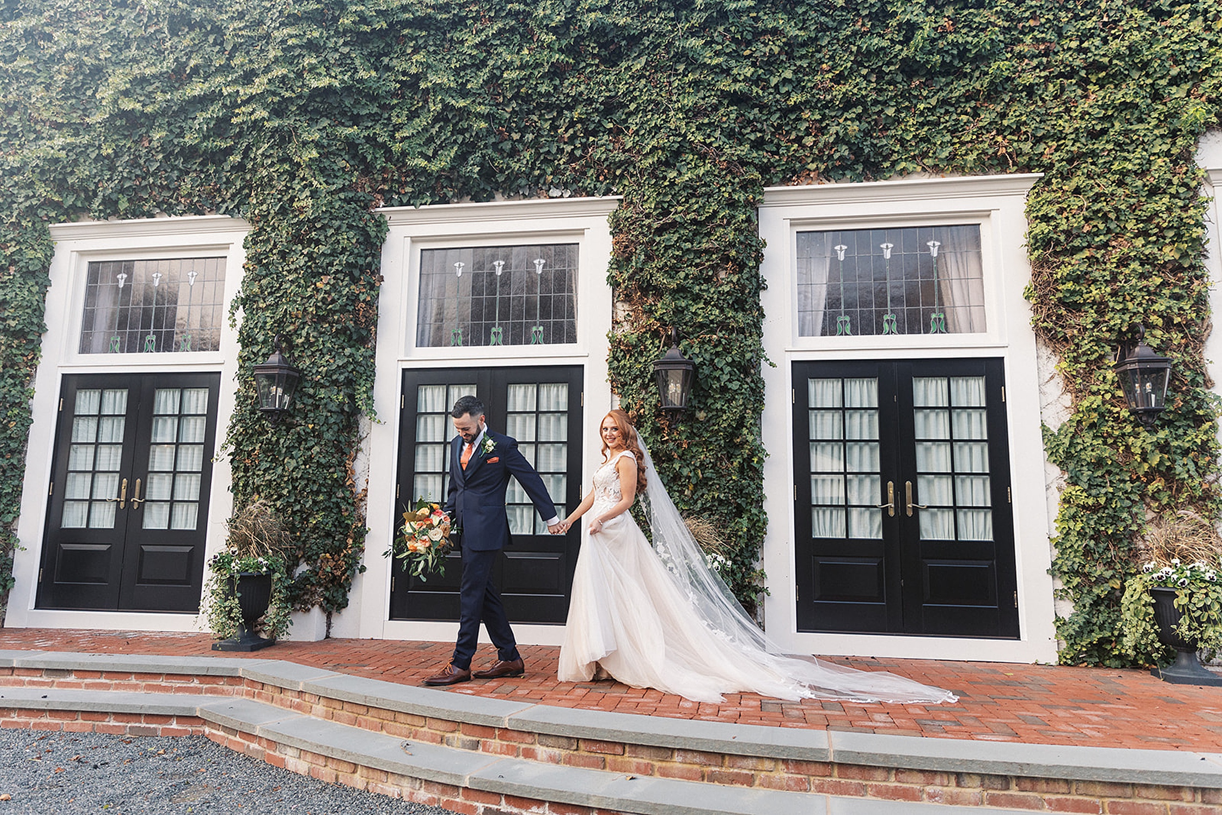 A groom leads his bride down a brick path in front of an ivy covered building at a David’s Country Inn Wedding