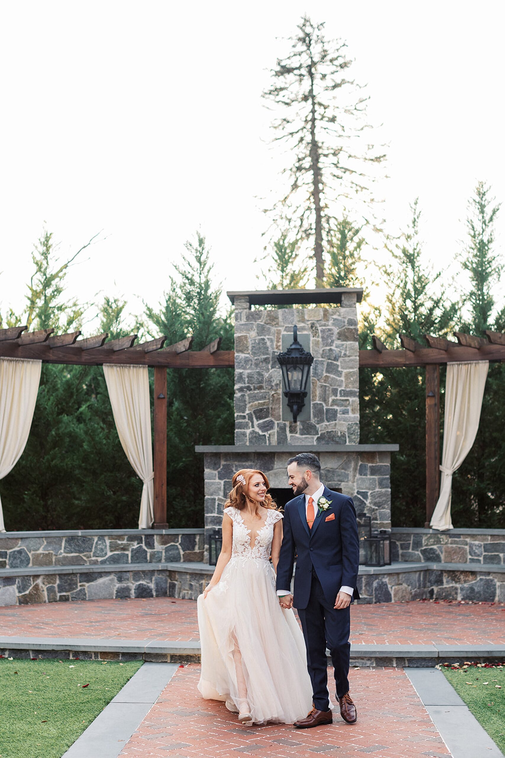 A bride and groom smile at each other while walking down a brick path past a stone fireplace