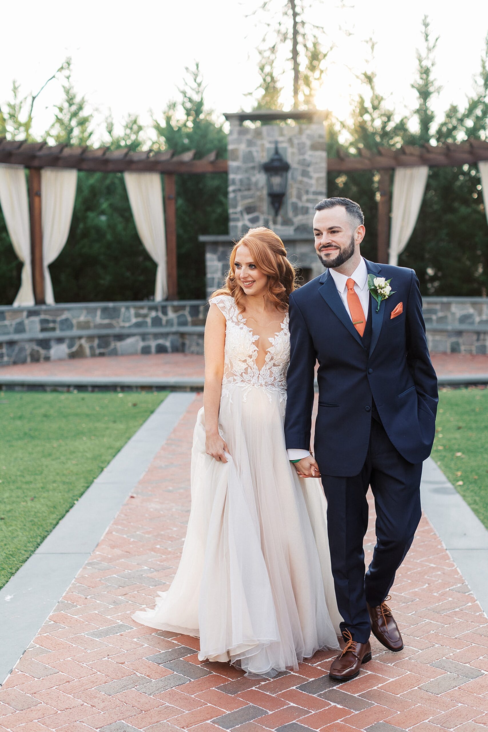 Newlyweds hold hands walking down a brick path in a garden at sunset