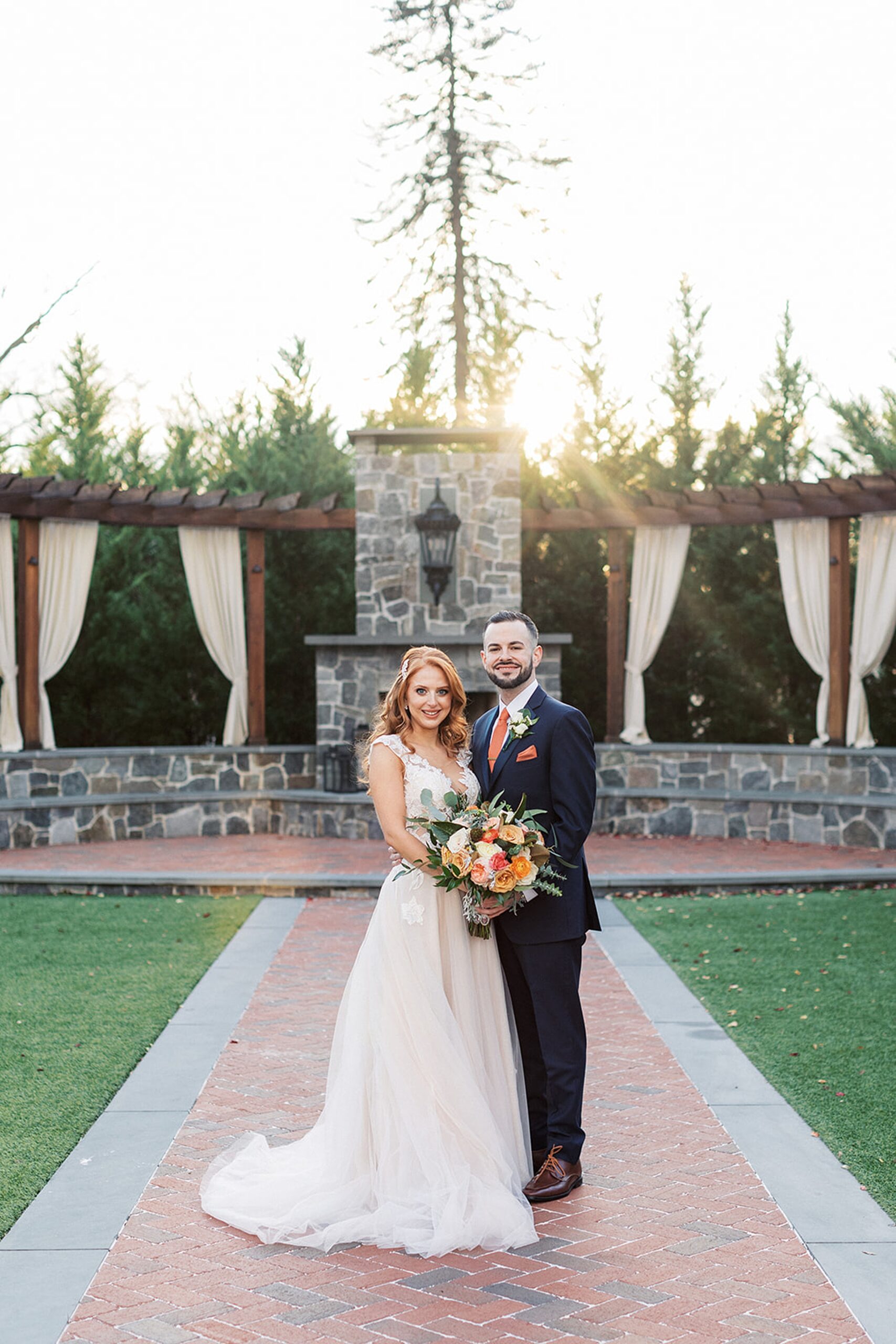 Newlyweds stand outside in a garden on a brick path at sunset holding the large orange bouquet at a David's country inn wedding