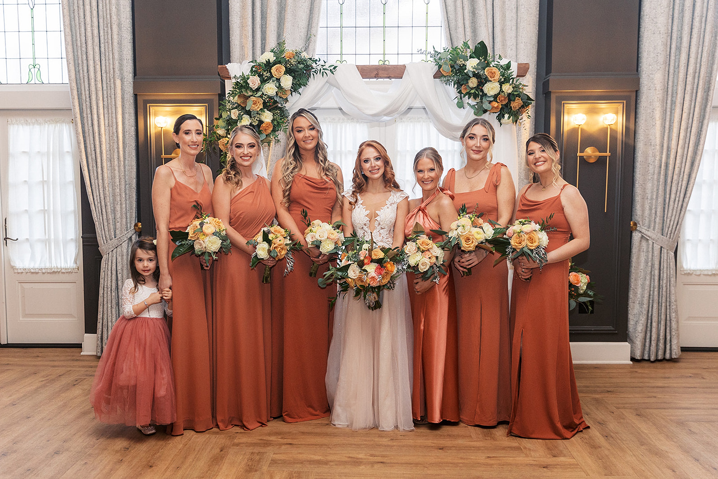 A bride stands under her ceremony arbor holding a bright bouquet with her bridesmaids and flower girl in matching orange dresses
