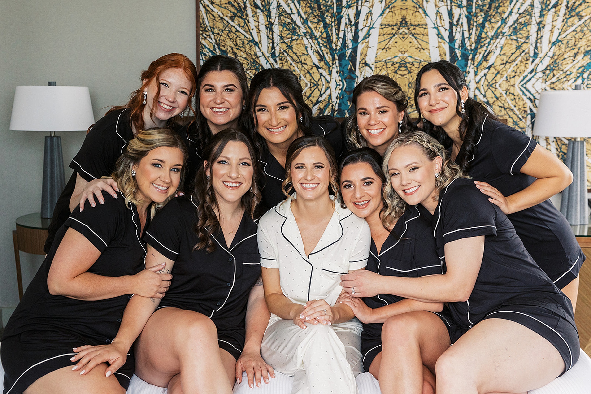A bride sits in white pajamas with her bridal party surrounding her on a bed in black pajamas