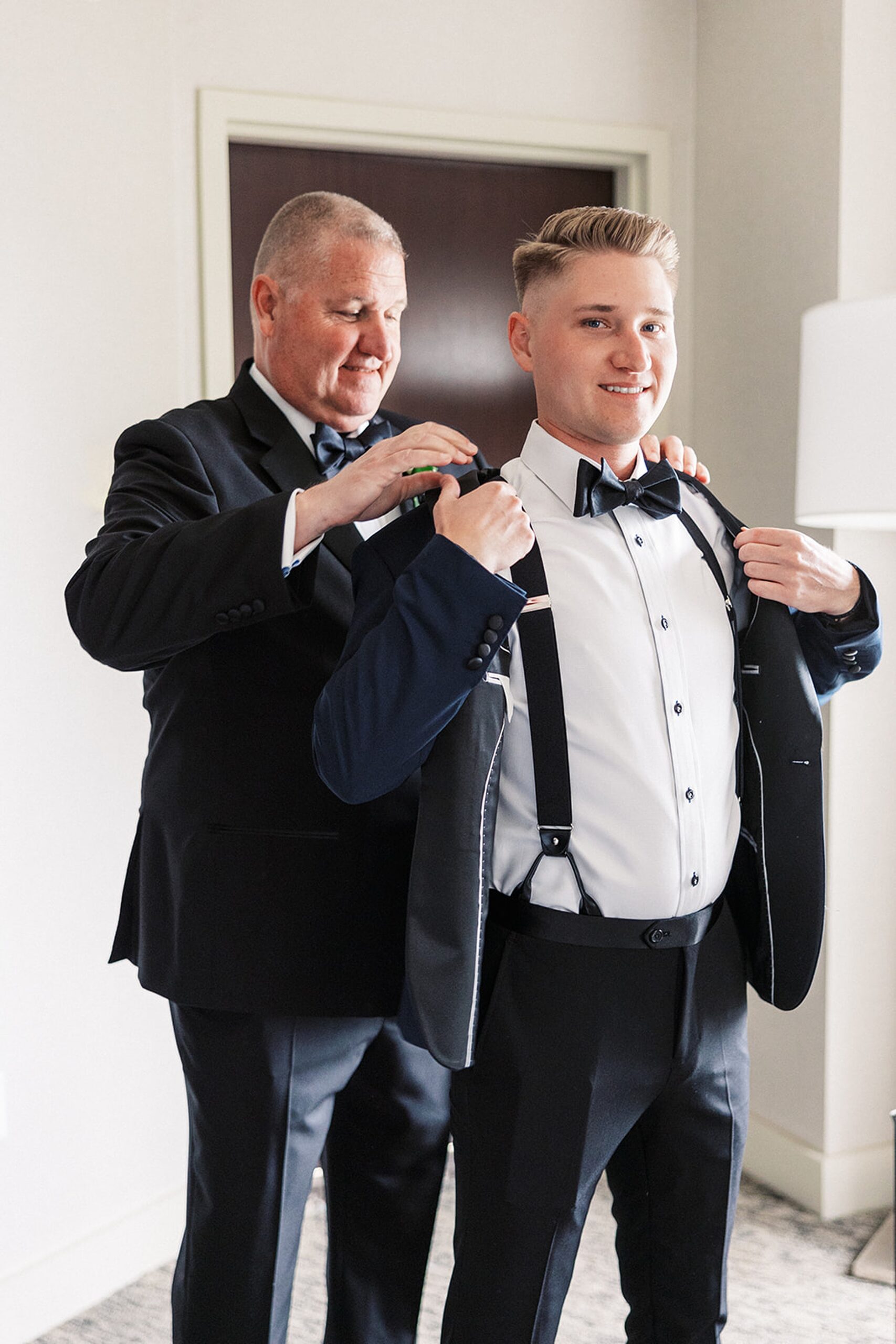 A father helps his son put on a tuxedo jacket while standing in a getting ready room