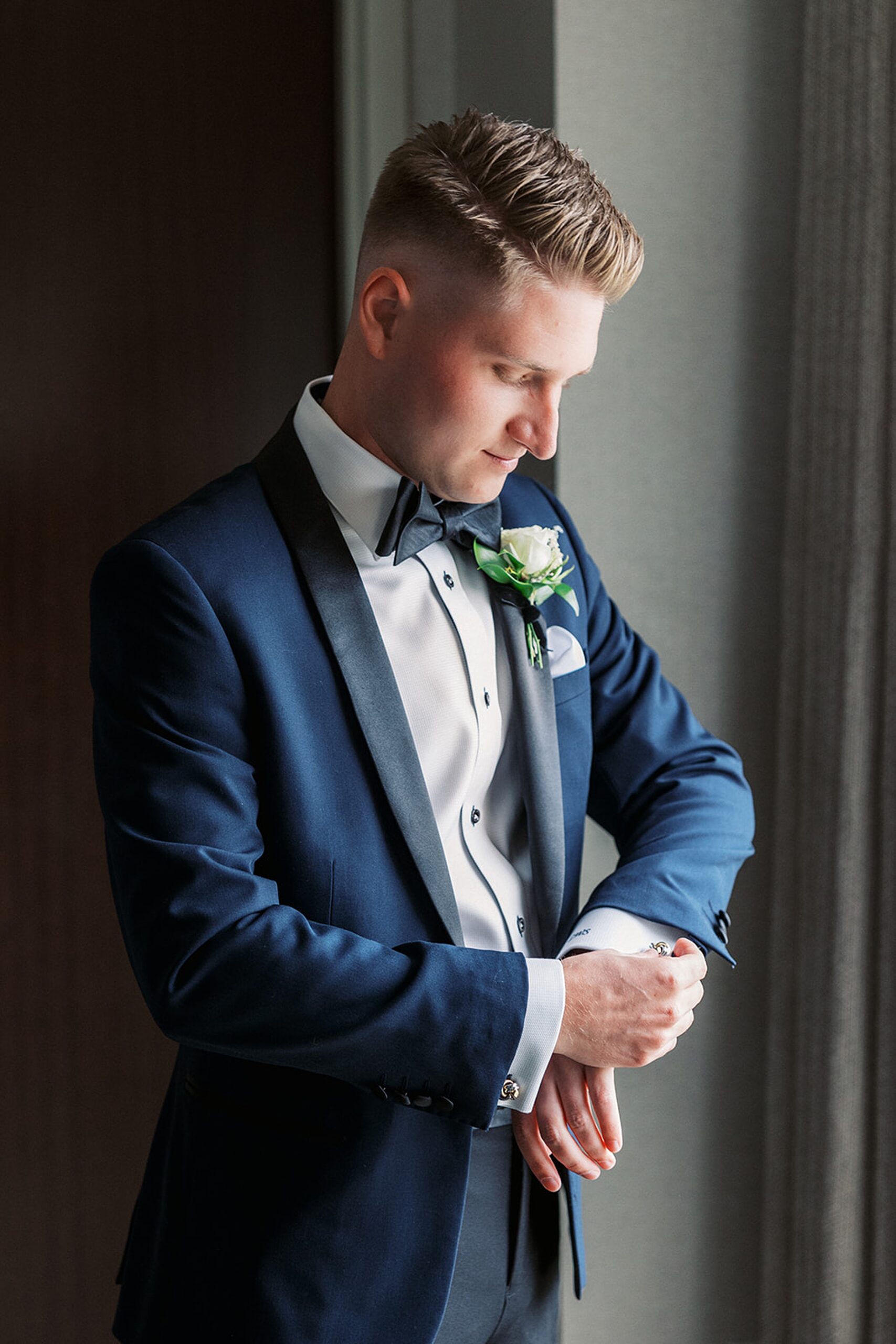 A groom stands in a window adjusting his cufflinks