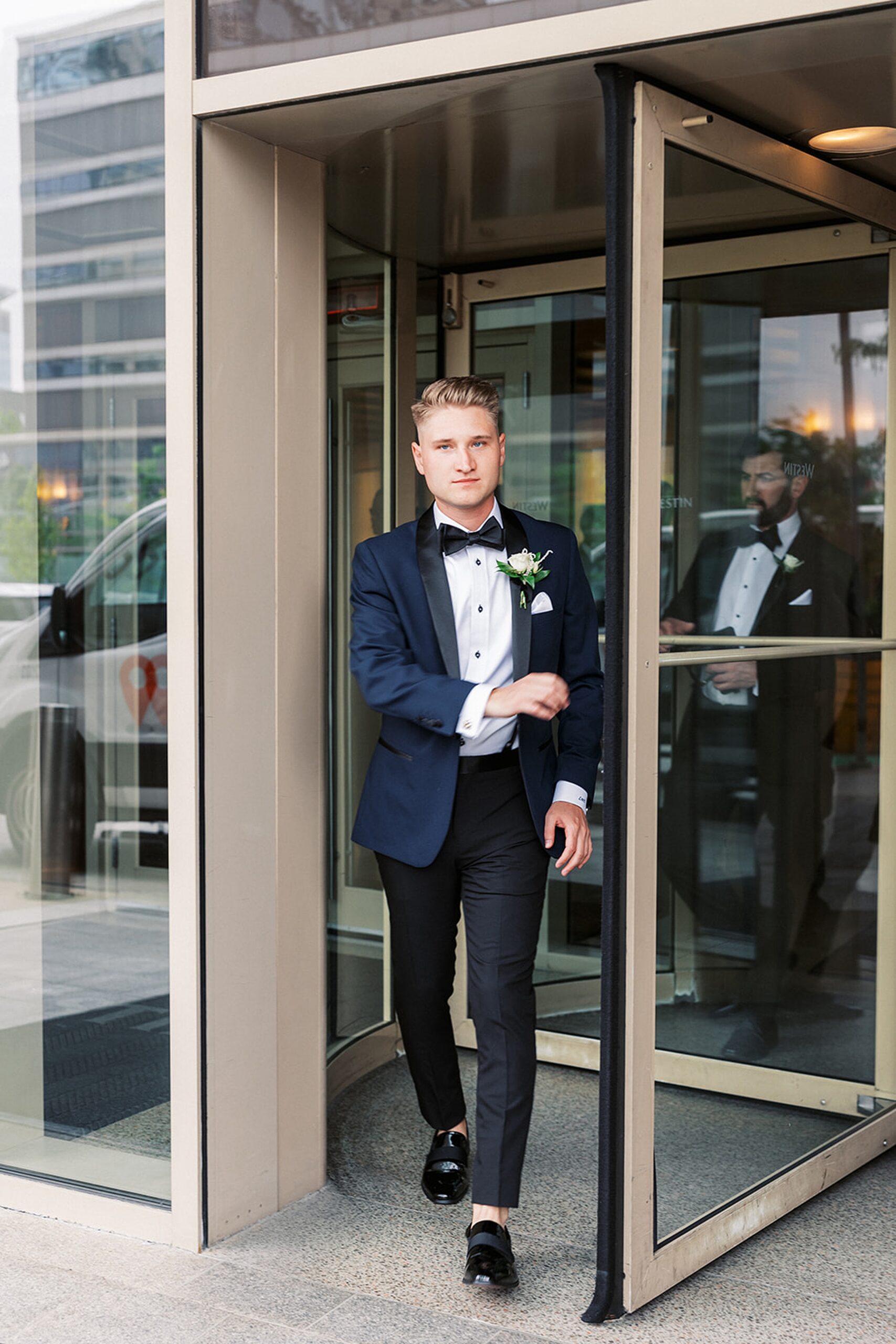 A groom walks through a revolving door in a blue tuxedo jacket and black pants