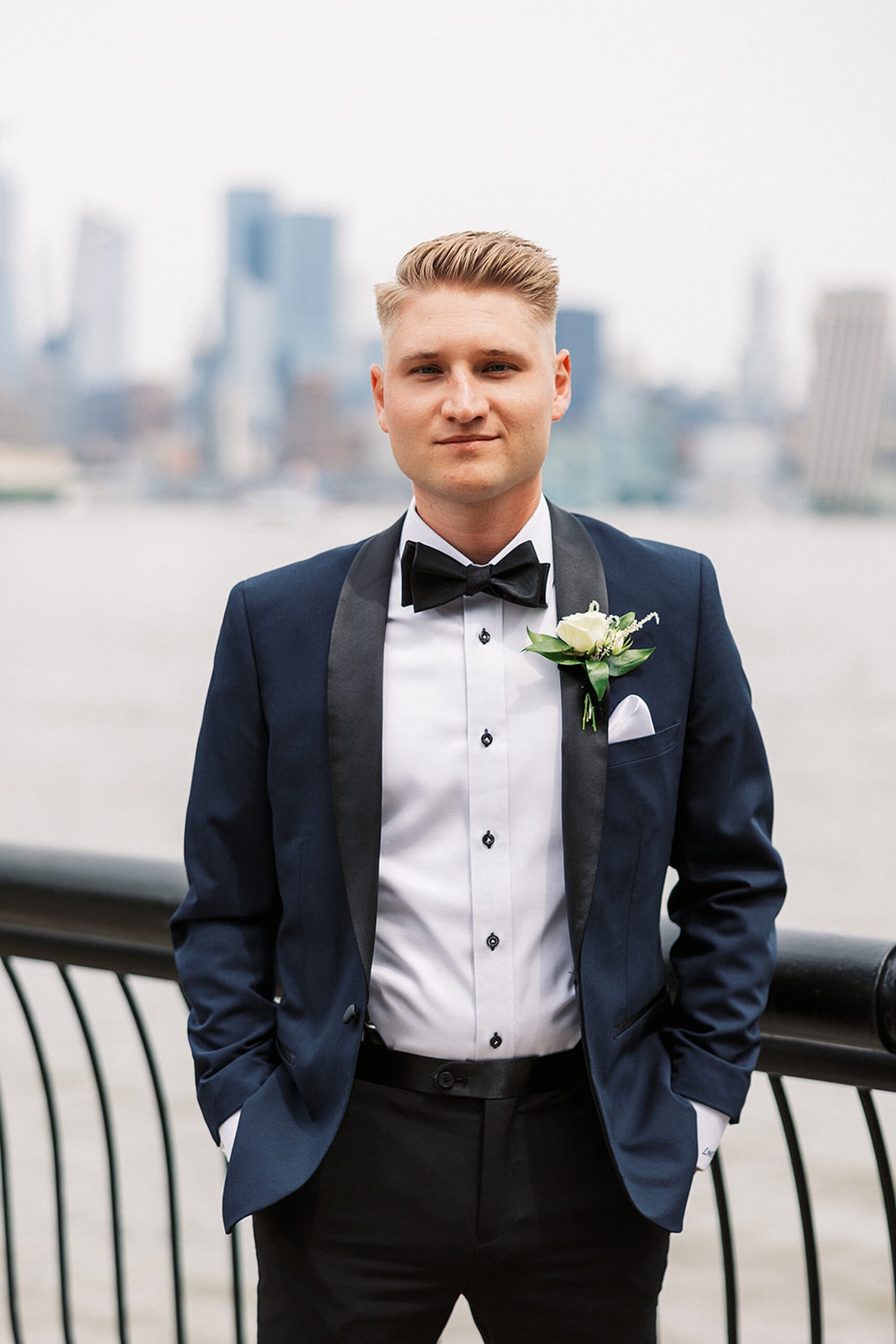 A groom in a blue tux stands on a riverfront boardwalk with hands in his pockets