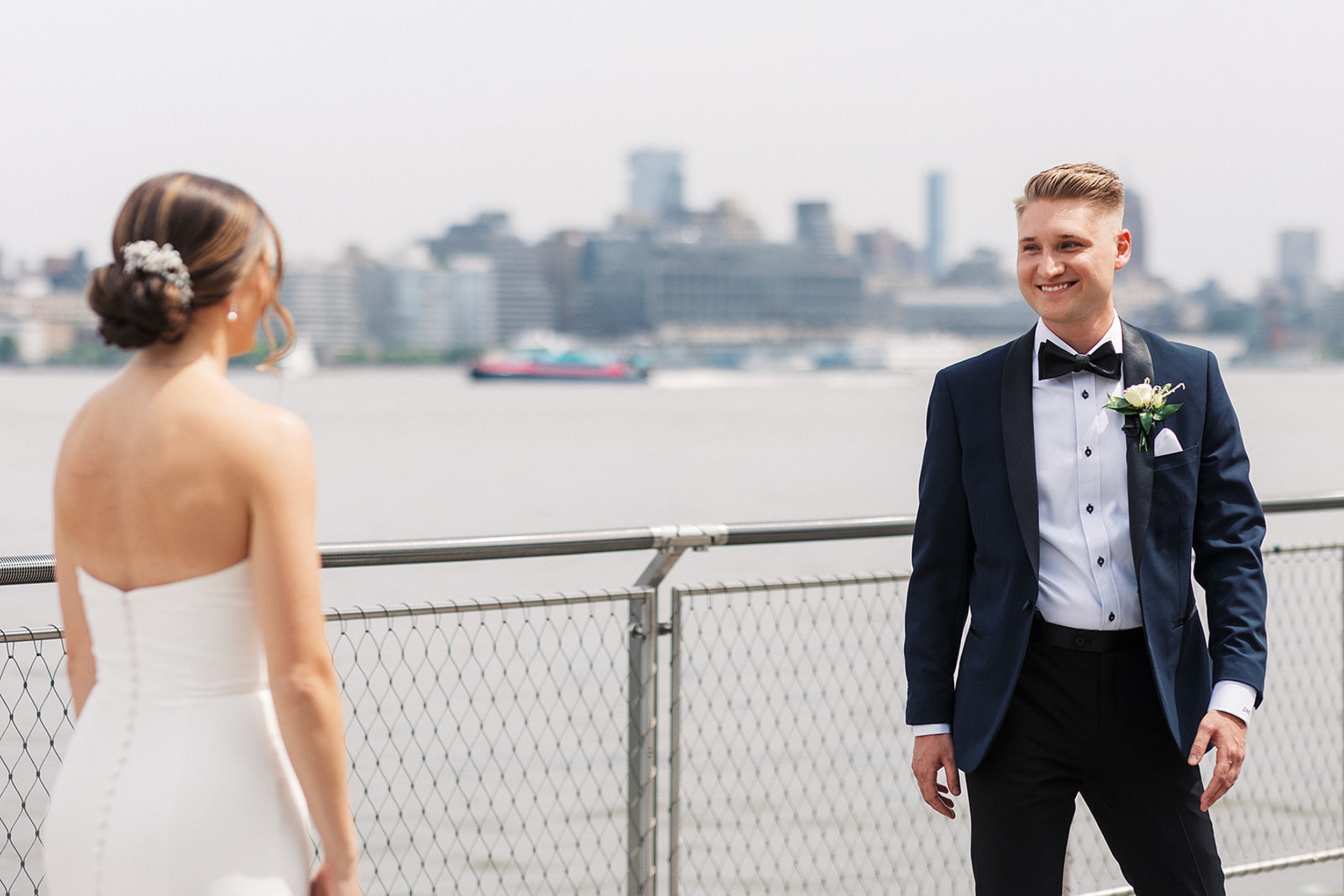 A groom smiles big as his bride walks toward him on a waterfront boardwalk