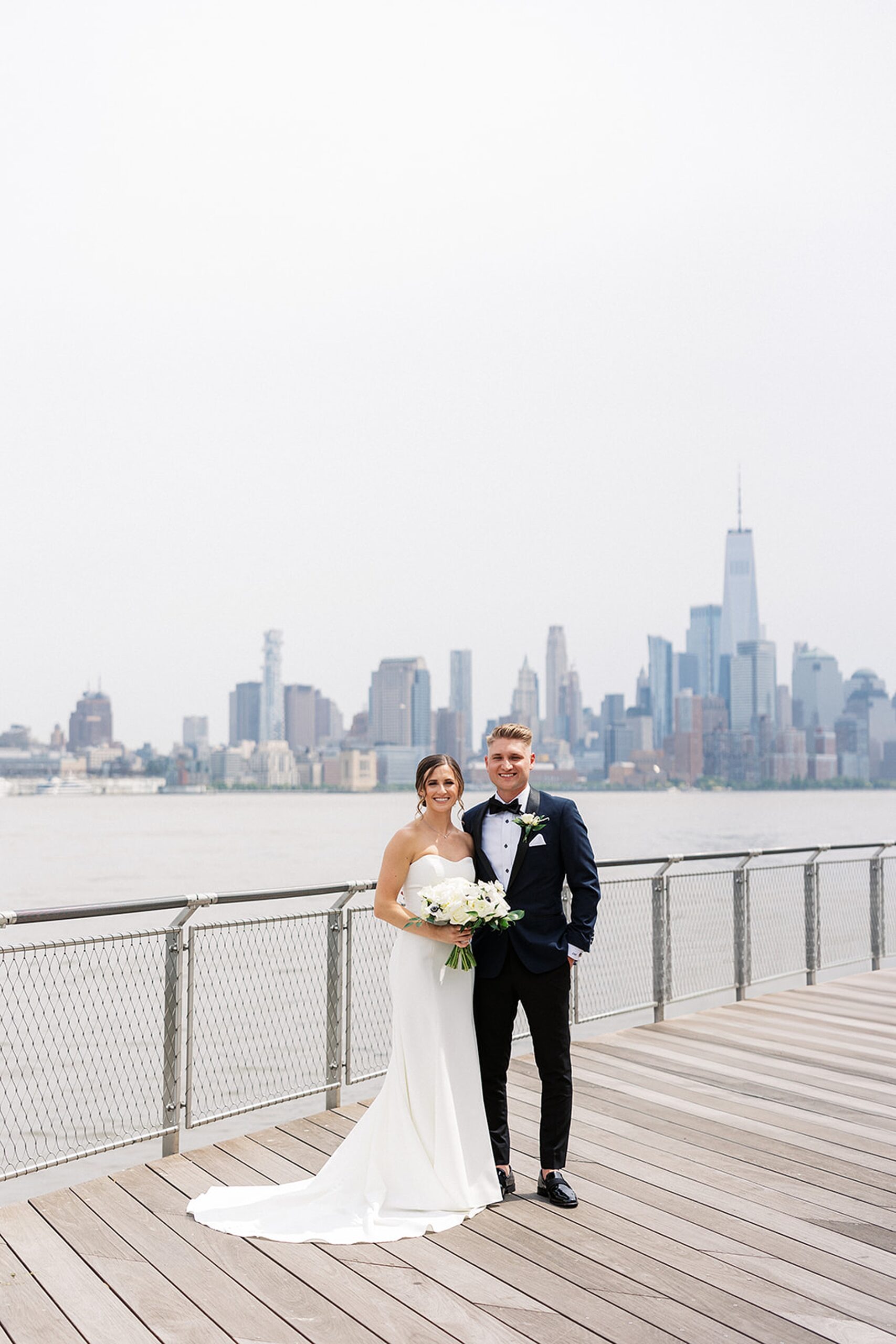 Newlyweds stand on a boardwalk in front of the Manhattan skyline