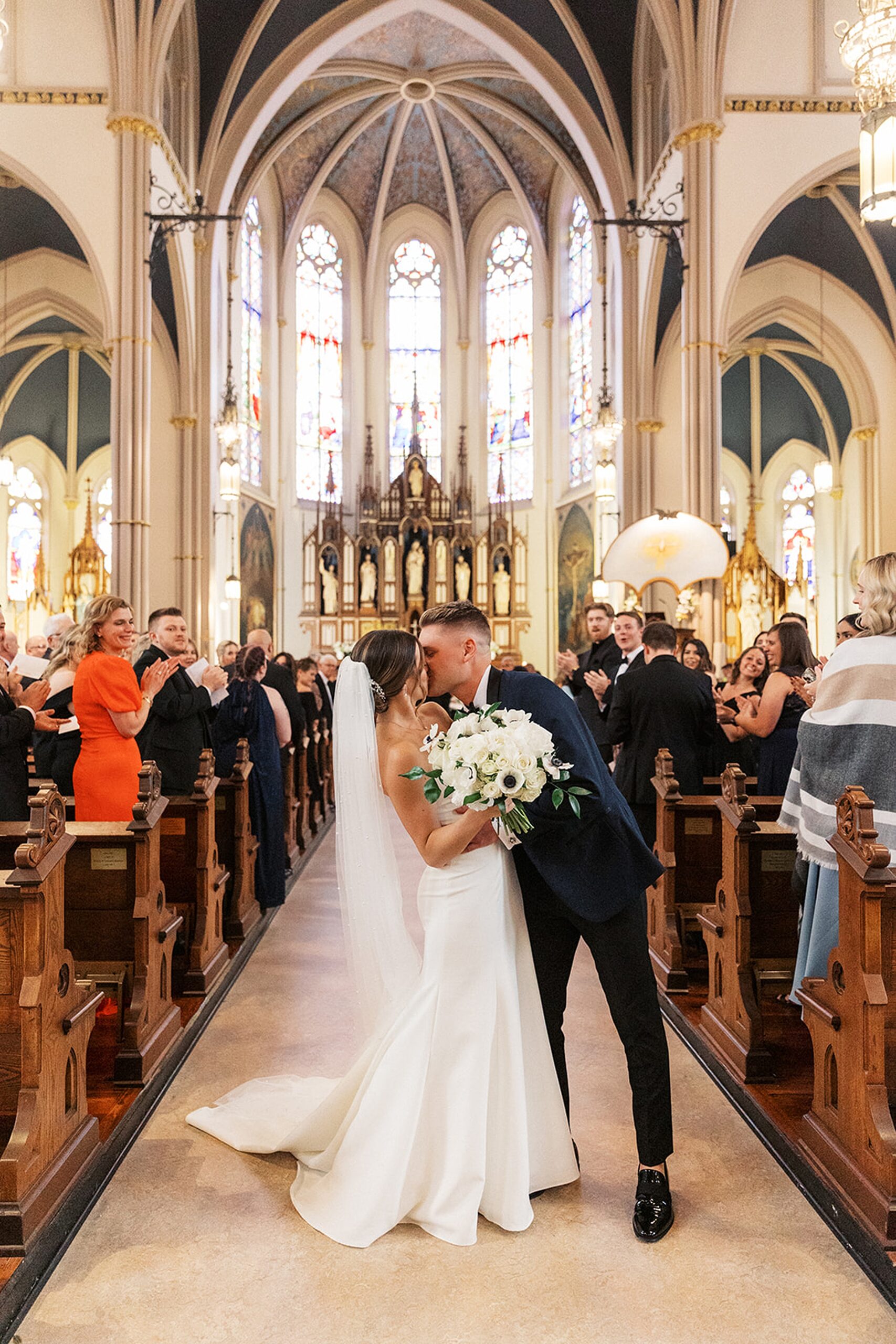 Newlyweds kiss at the end of the aisle in a large church while their guests clap and cheer