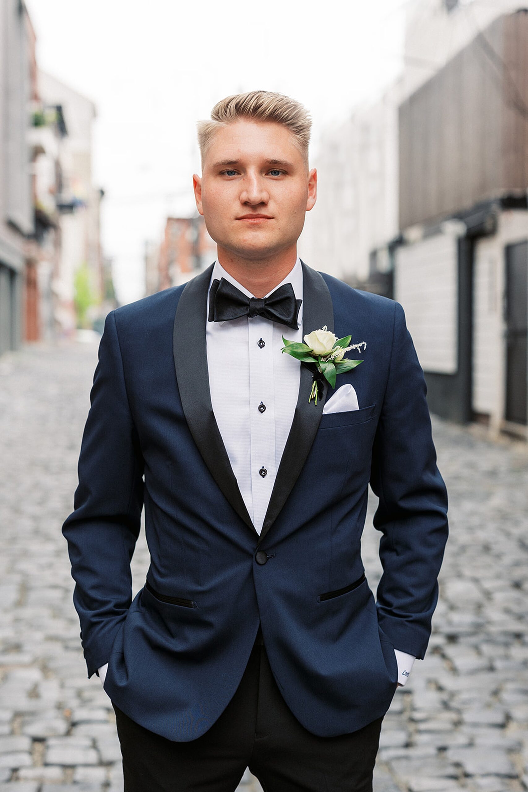 A groom stands in a stone paved alley with hands in his pockets in a blue tuxedo at a Hudson House wedding