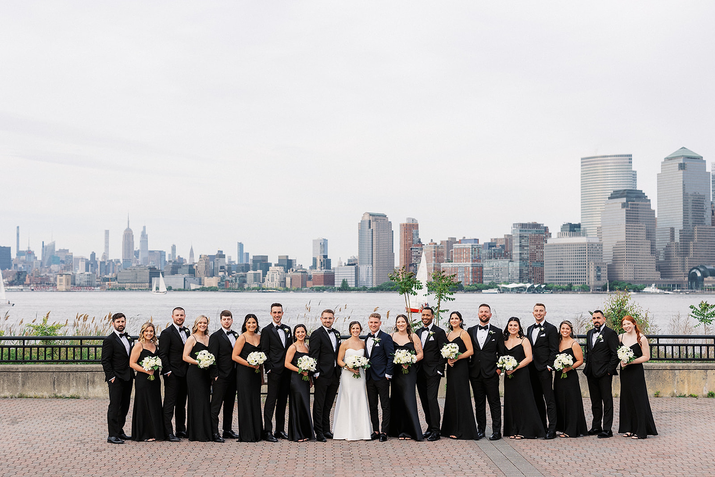 A wedding party stands together in a park on the river with the New York City skyline behind them