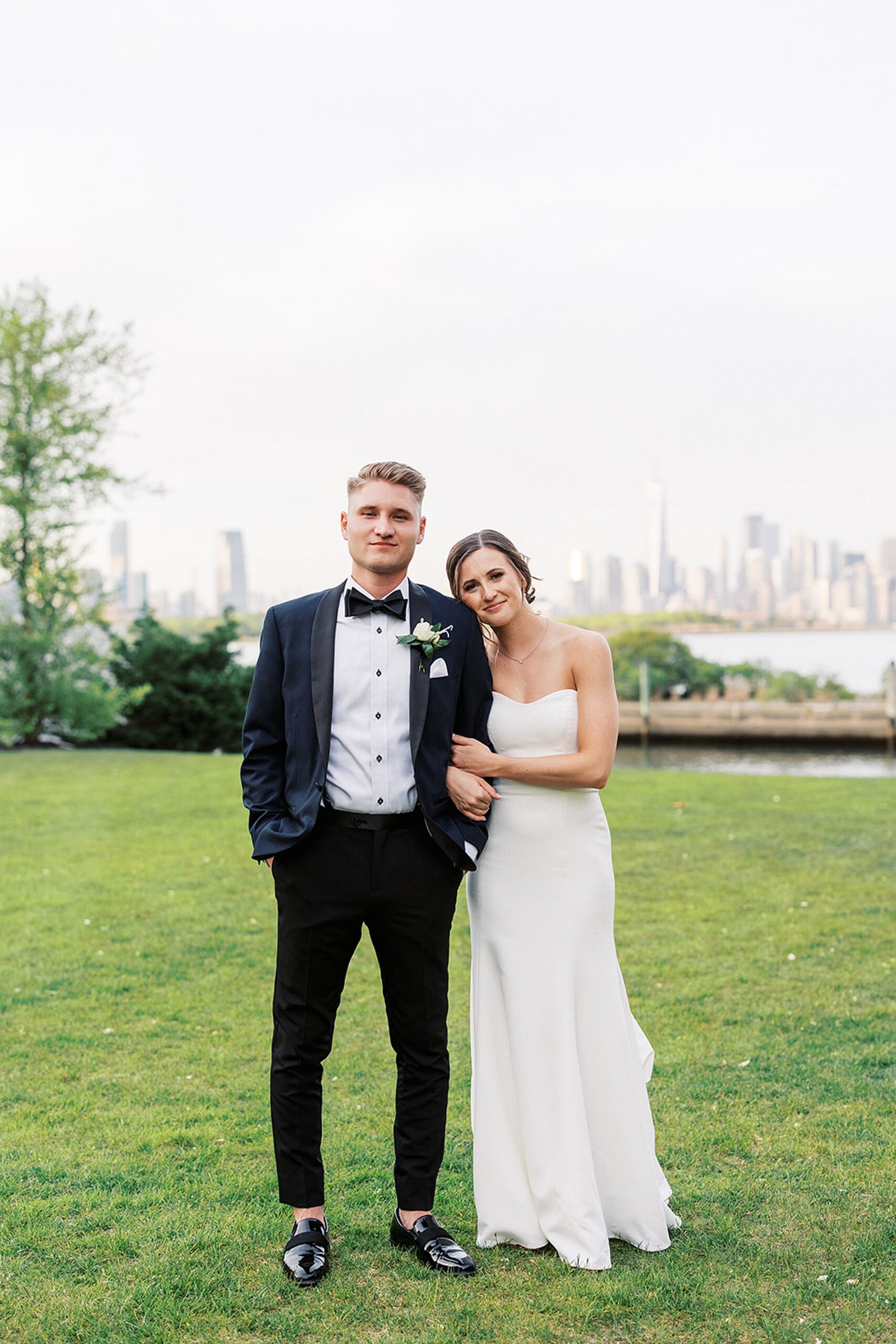 A bride holds onto the arm of her husband in a grassy park with Manhattan in the background