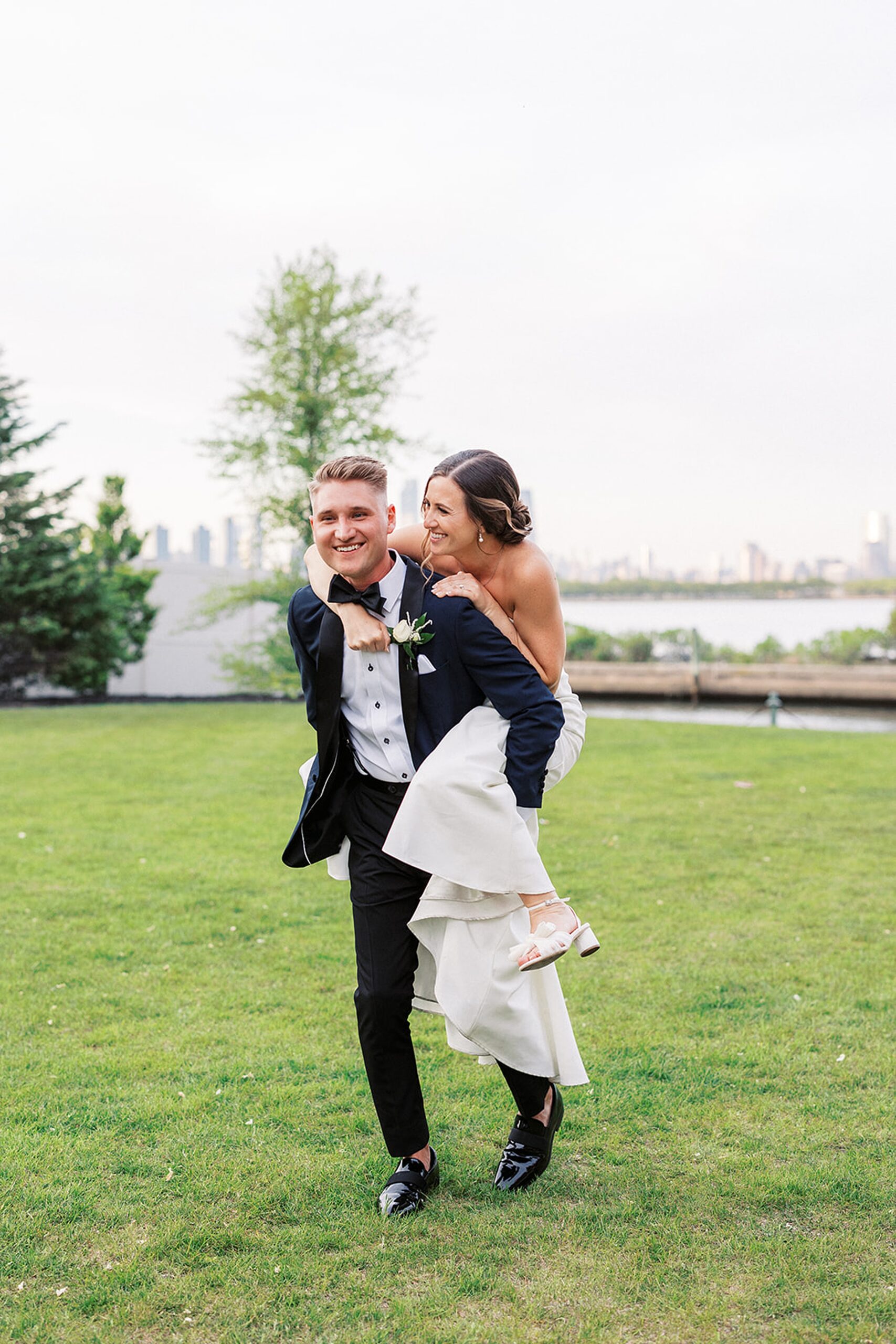 A bride hops on her husbands back while they walk through a grassy field