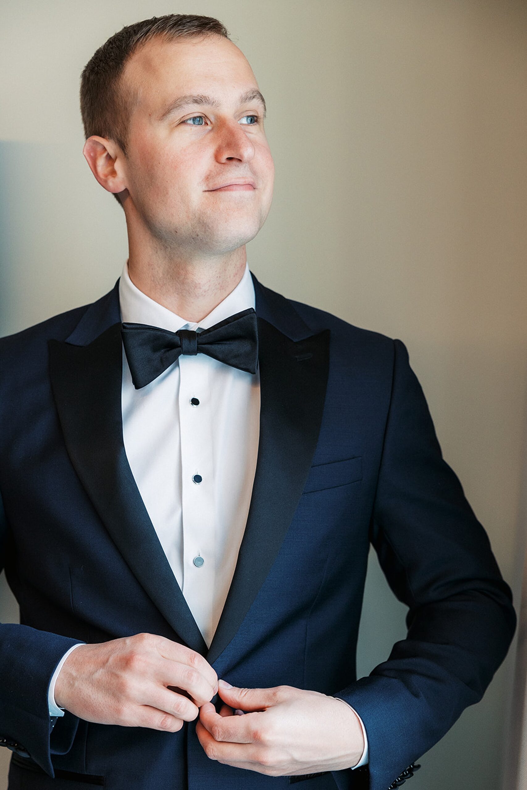 A groom buttons his black jacket while looking out a window