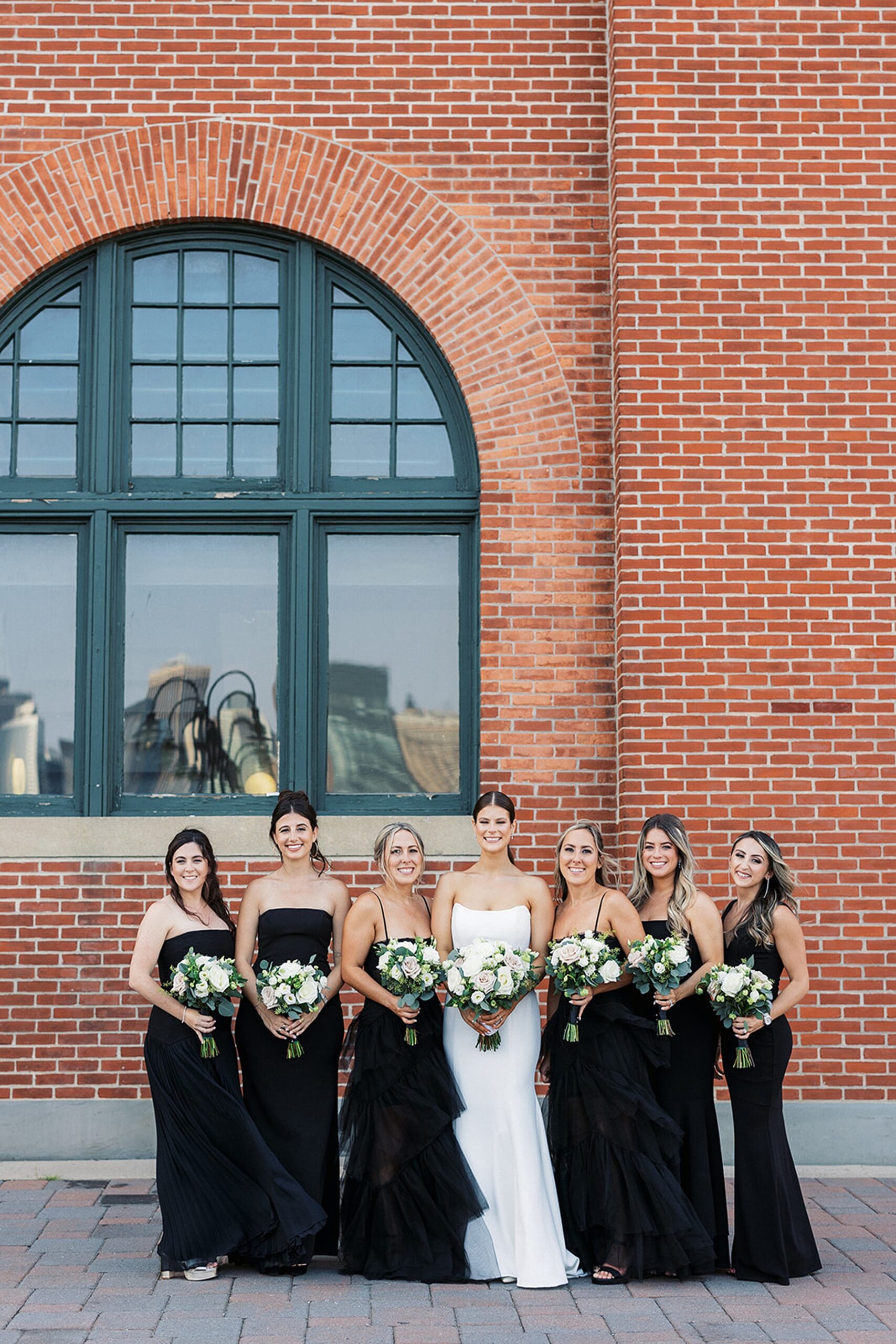 A bride stands by a brick building with her six bridesmaids