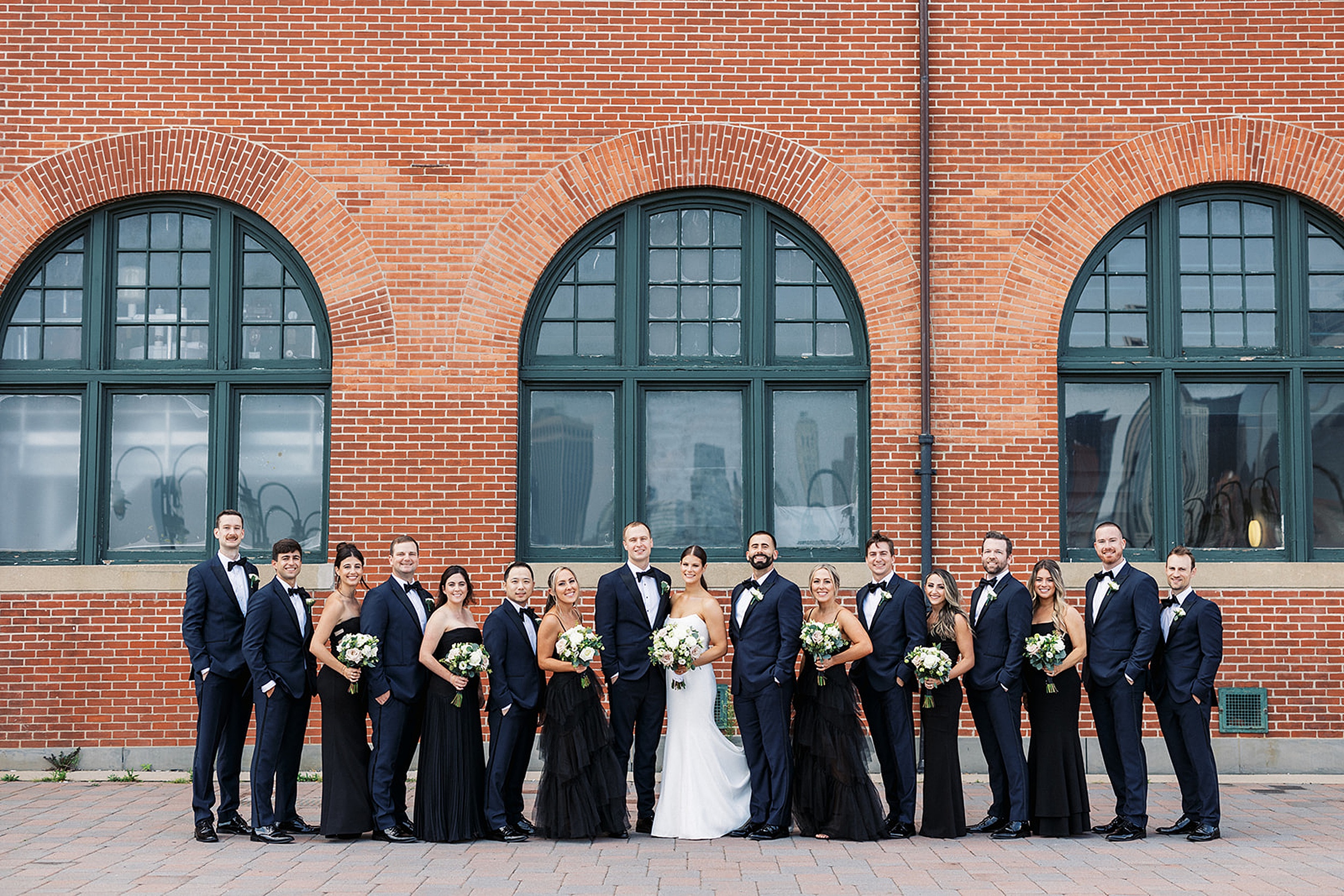 A bride and groom stand in the middle of their large wedding party by a brick building
