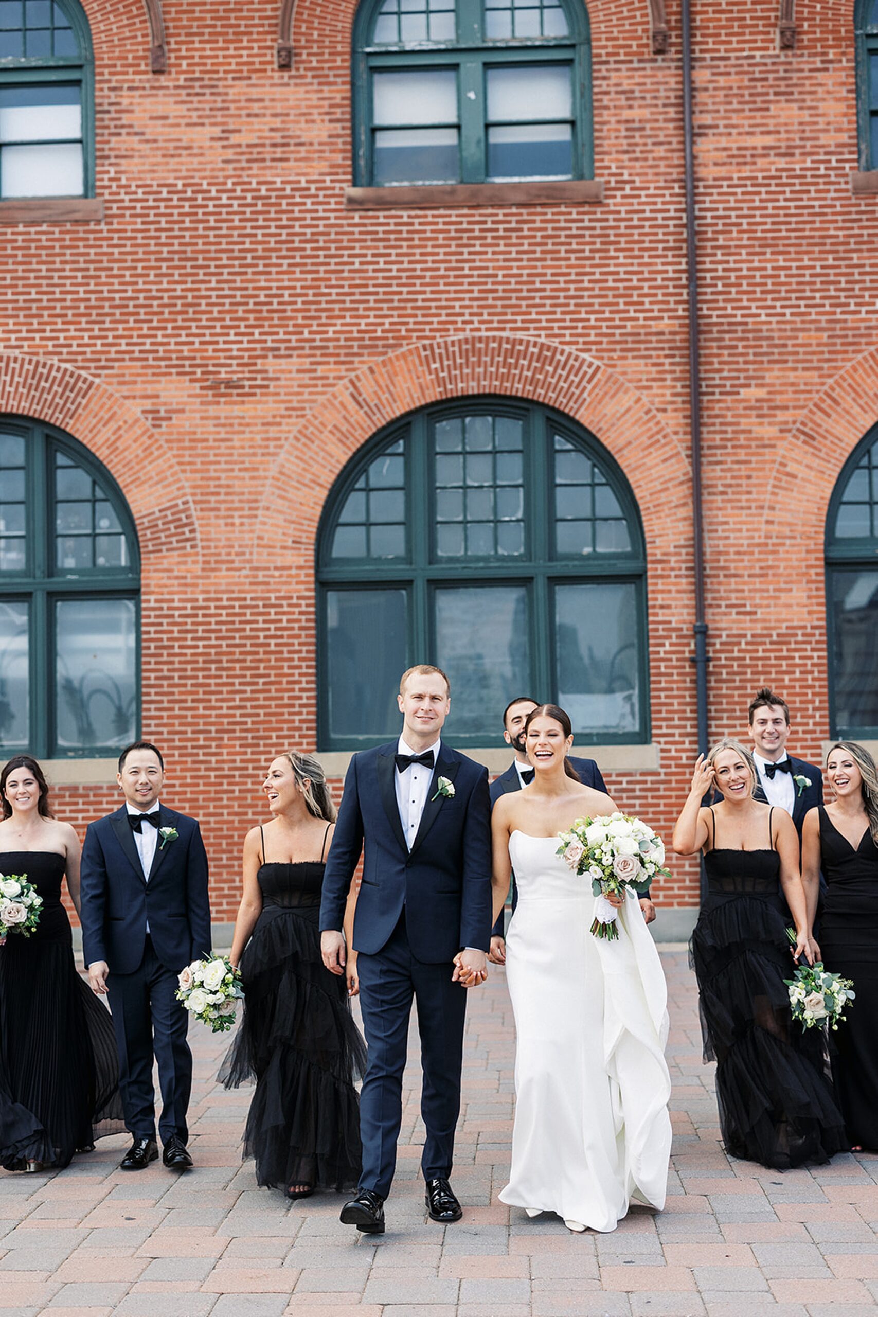 Newlyweds hold hands while walking down a sidewalk surrounded by their wedding party