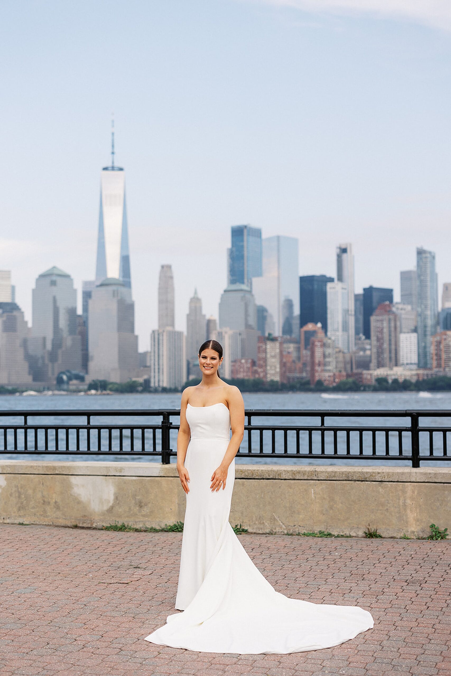 A bride stands in a brick paved riverfront park with Manhattan behind her