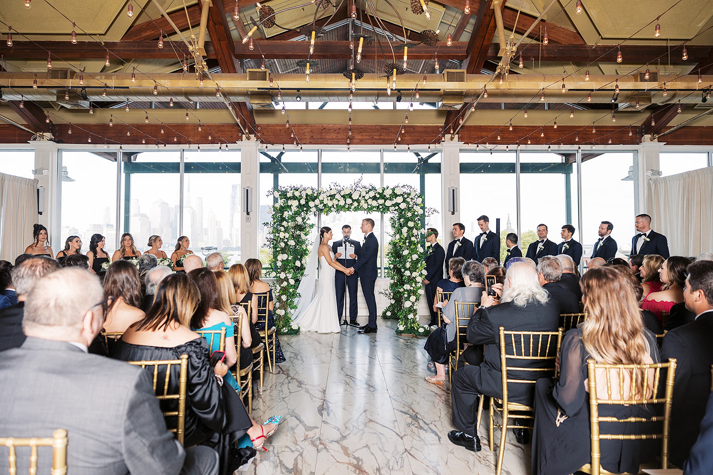 Newlyweds hold hands under an arch of white roses during their wedding ceremony at Liberty House Wedding venue