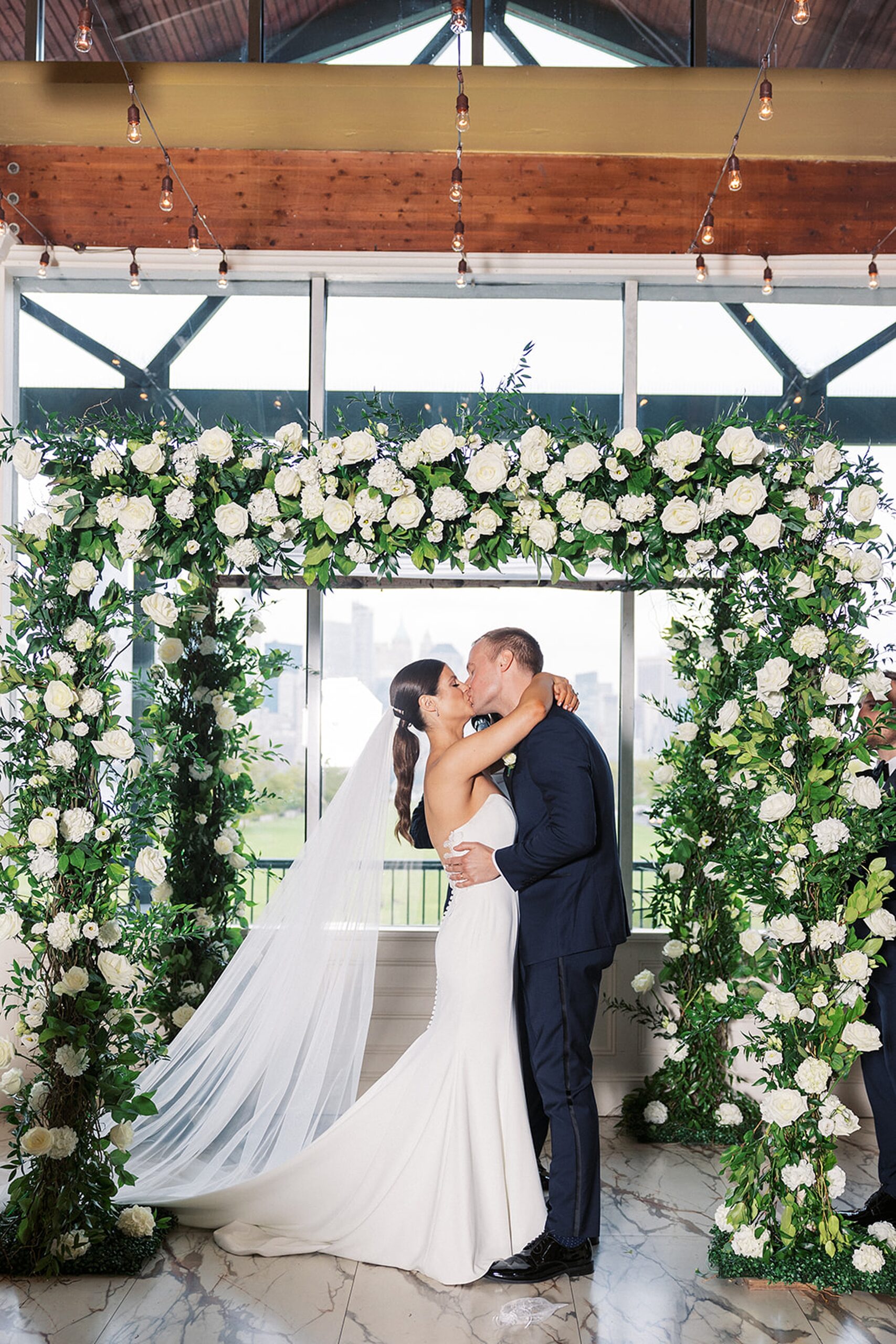 Newlyweds kiss at the end of their wedding ceremony under a white rose flower arch