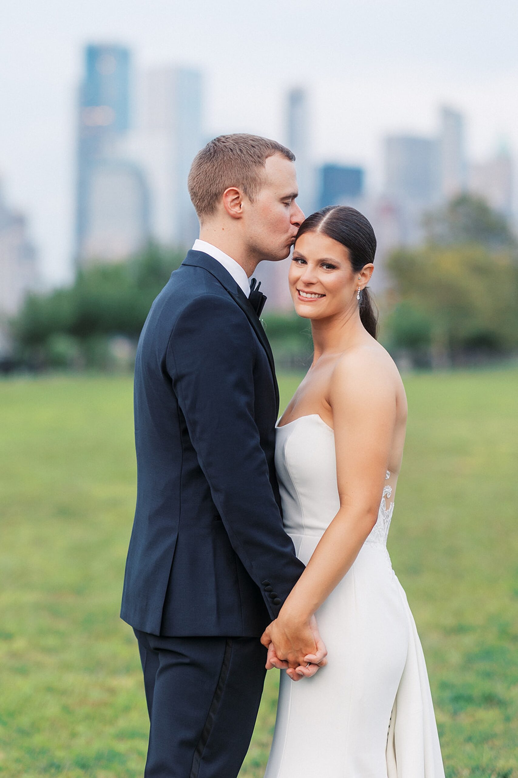 A groom kisses his bride on the temple while holding hands and standing in a grassy field