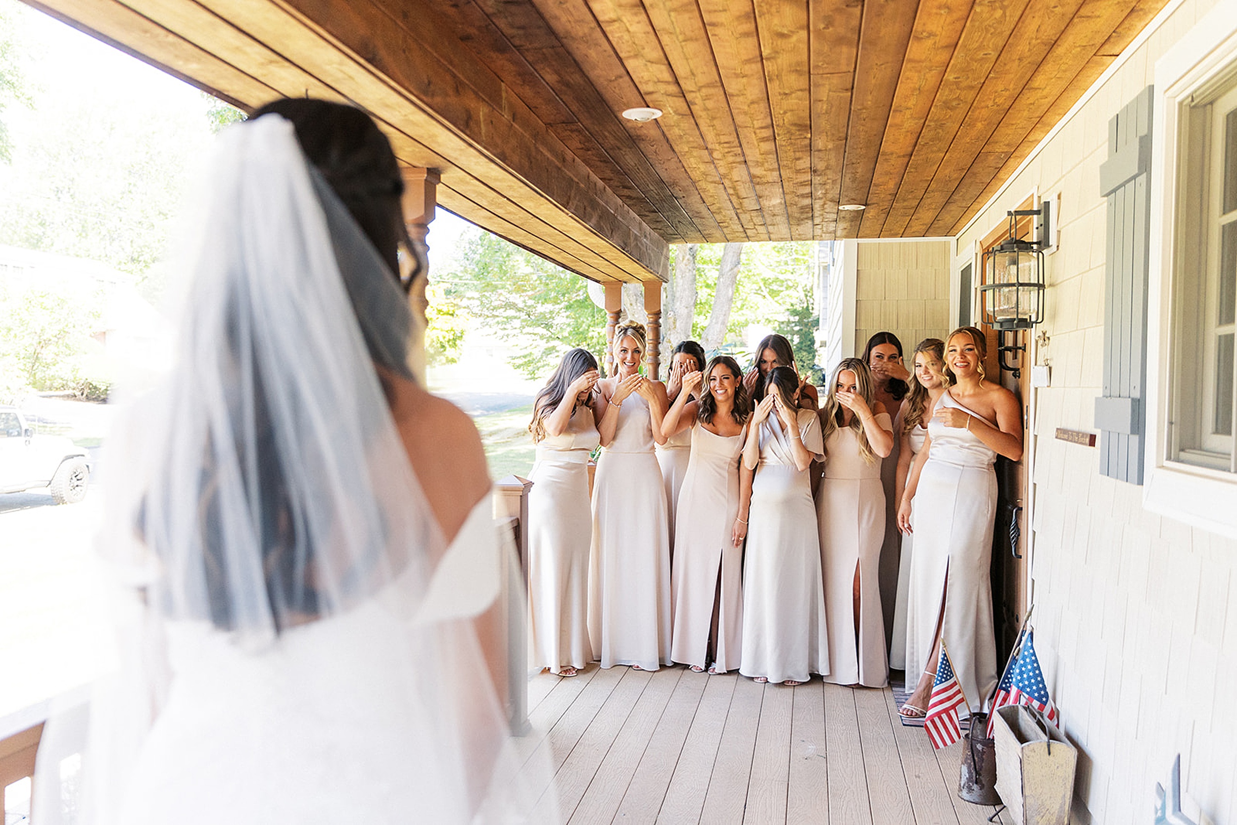 A bride stands on a porch while her bridesmaids see her for the first time and cheer for her