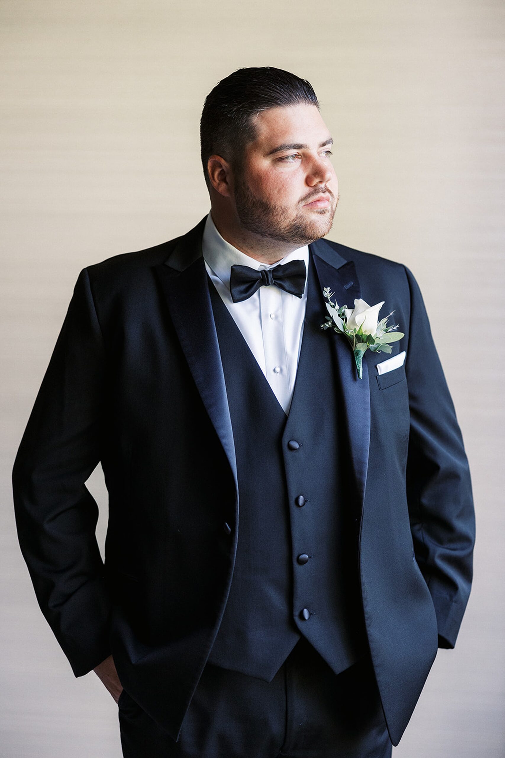 A groom stands with hands in his pockets while standing and looking out a window in a black tuxedo