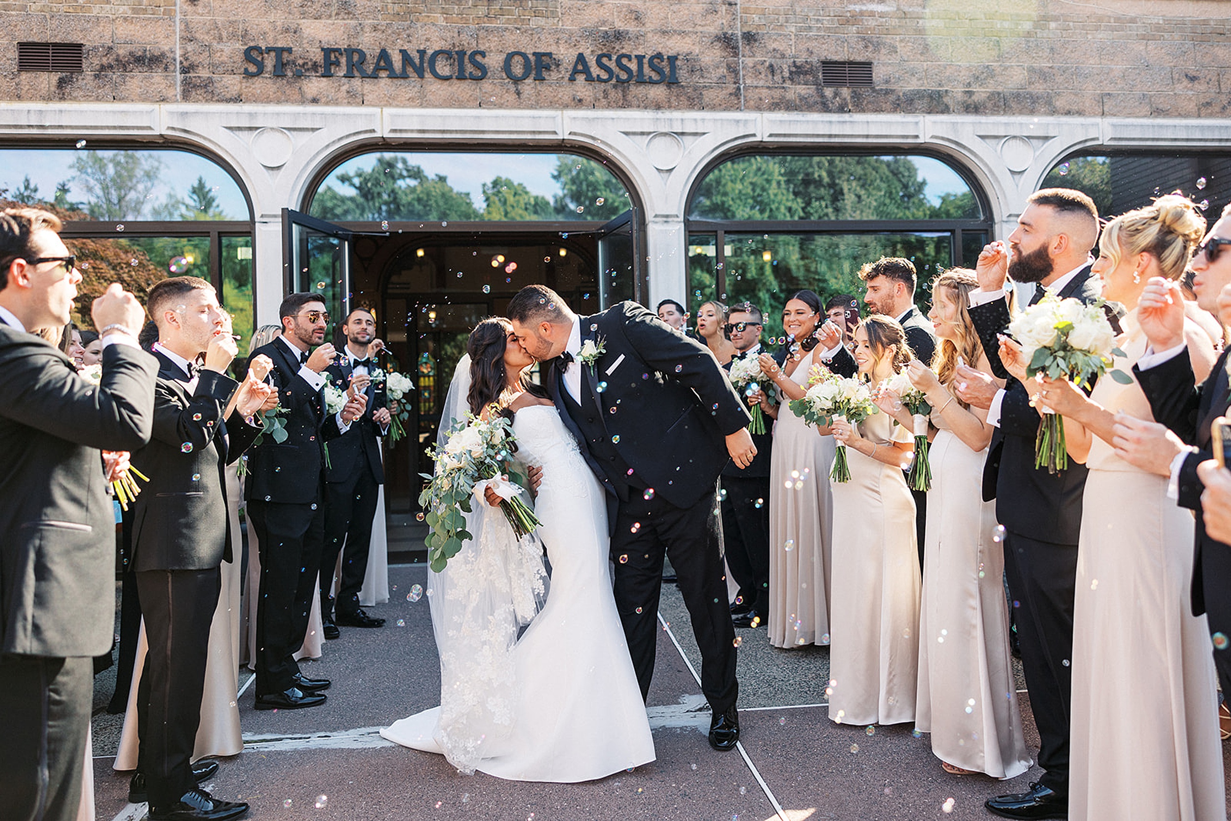 Newlyweds kiss and dip under a shower of bubbles surrounded by their wedding party