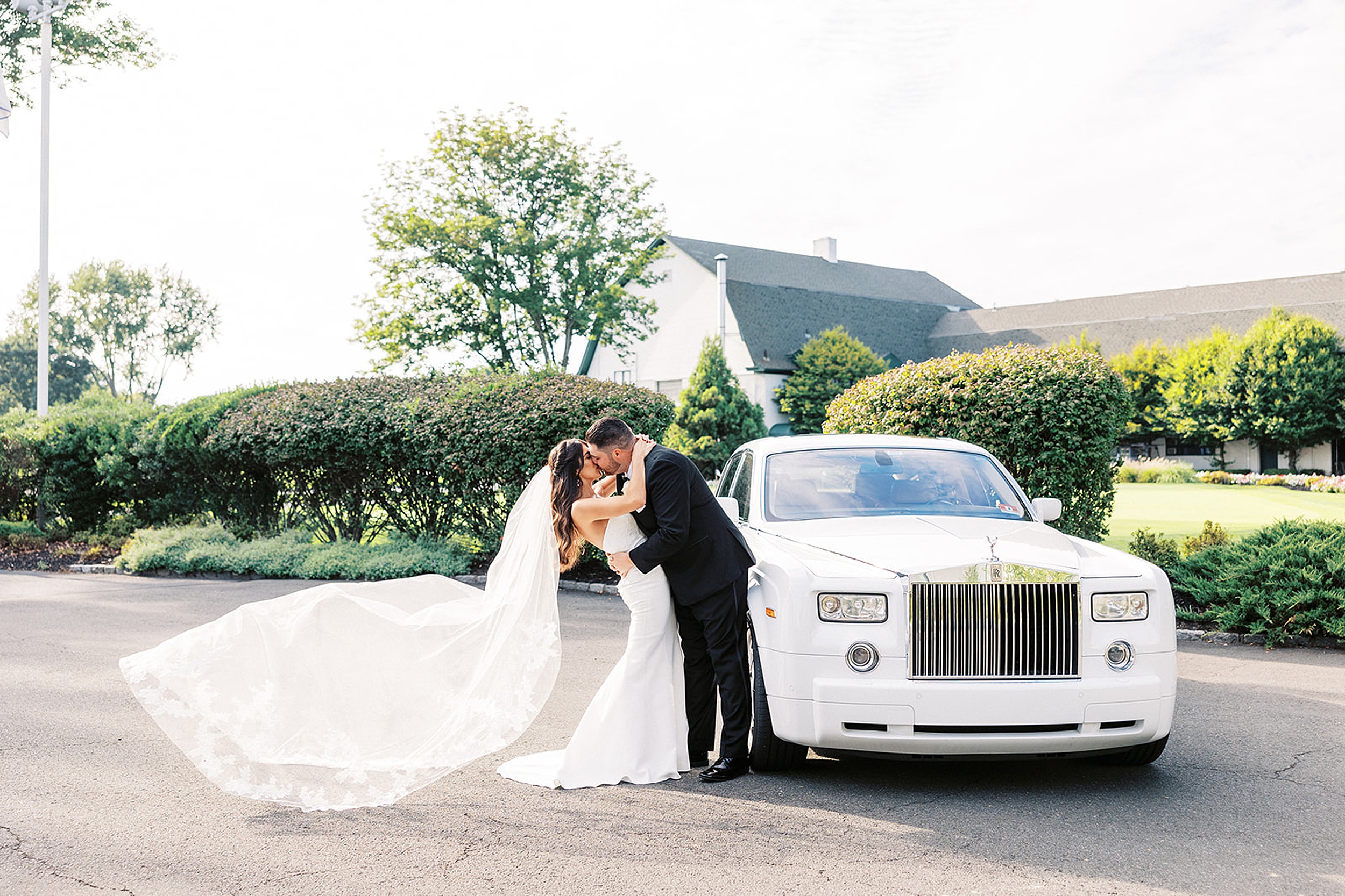Newlyweds kiss as the long veil dances behind them in the wind against a luxury White Rolls Royce
