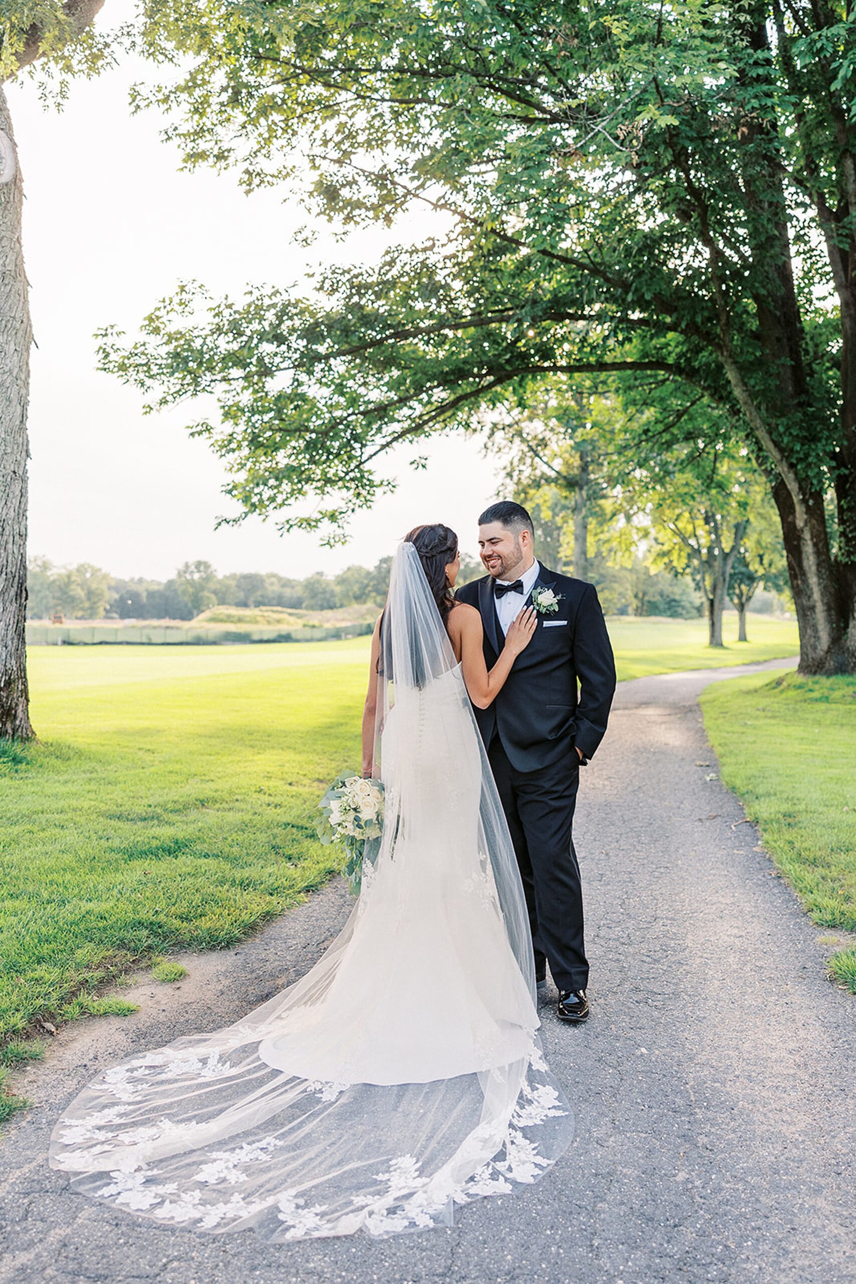 A bride in a long flowing veil and white dress rests her hand on her groom's chest while standing in a golf course path