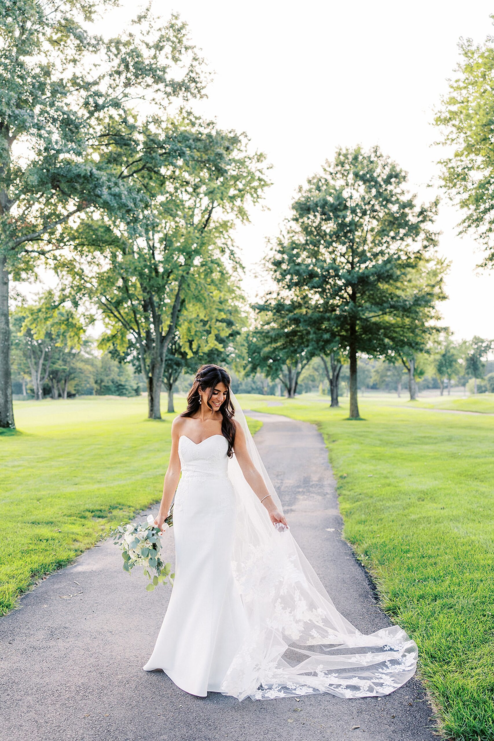 A bride plays with her ling veil and train in a golf course path at an edgewood country club wedding