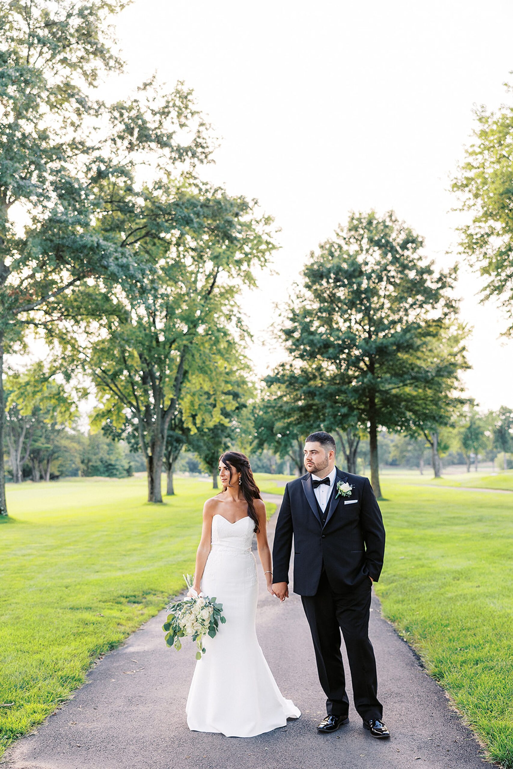 Newlyweds hold hands while walking down a paved walkway in a golf course