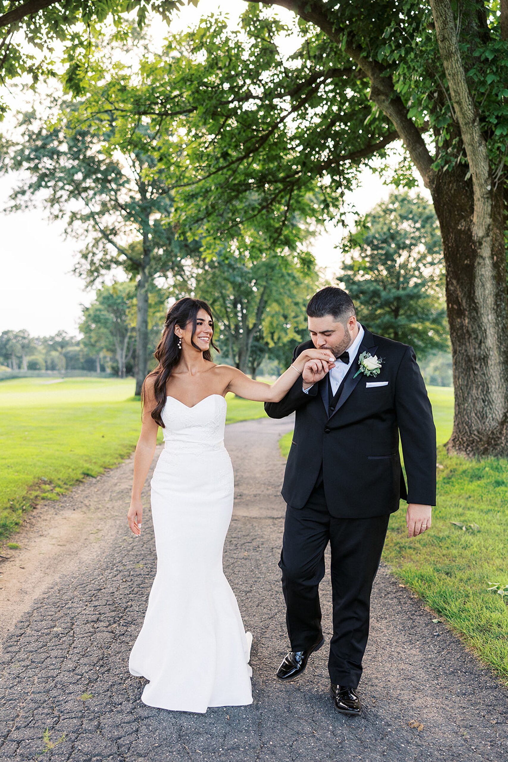 A groom kisses his bride's hand while they walk down a paved sidewalk under a large oak tree at an edgewood country club wedding