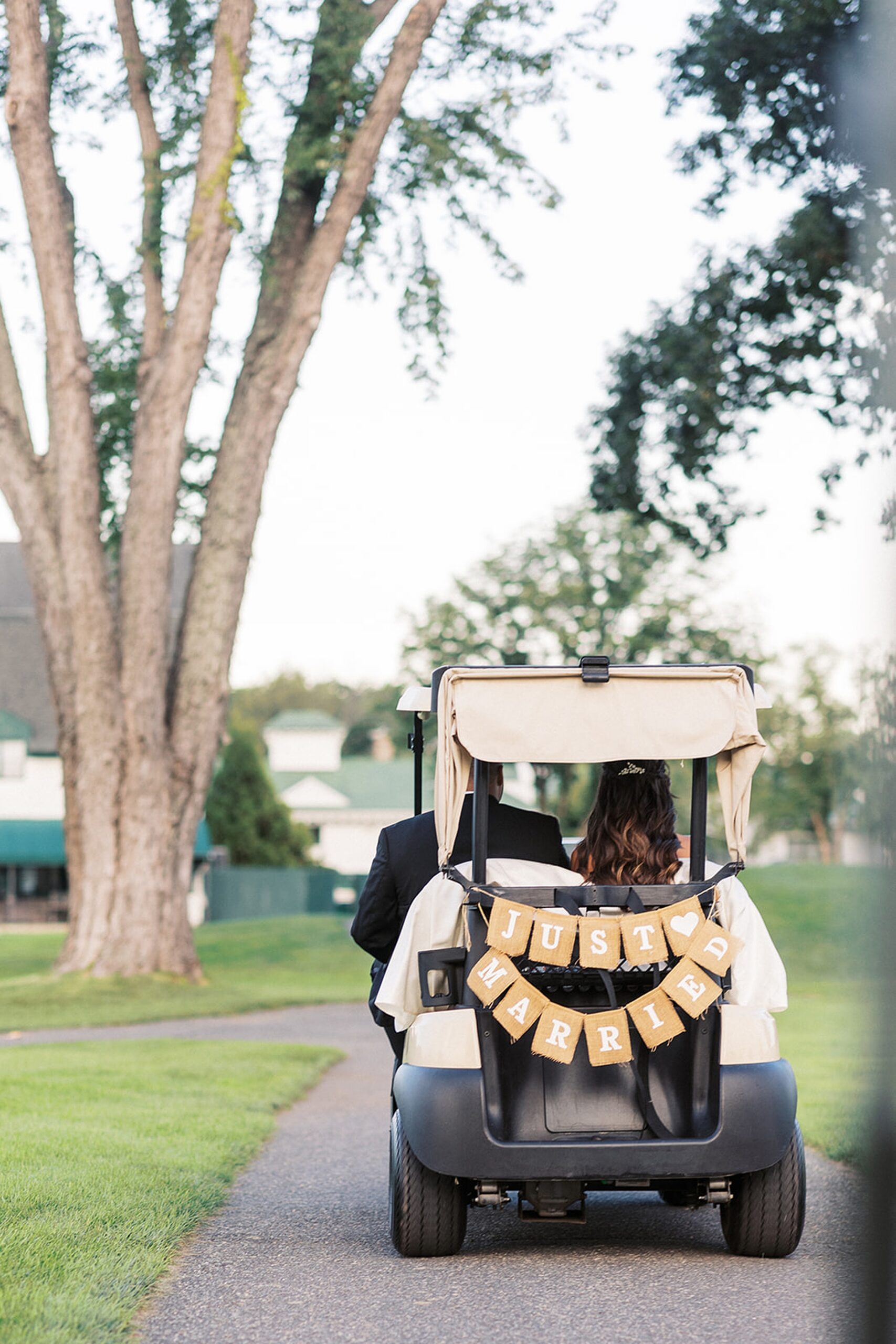 Newlyweds ride away in a golf cart with a "Just Married" banner on the back at an edgewood country club wedding