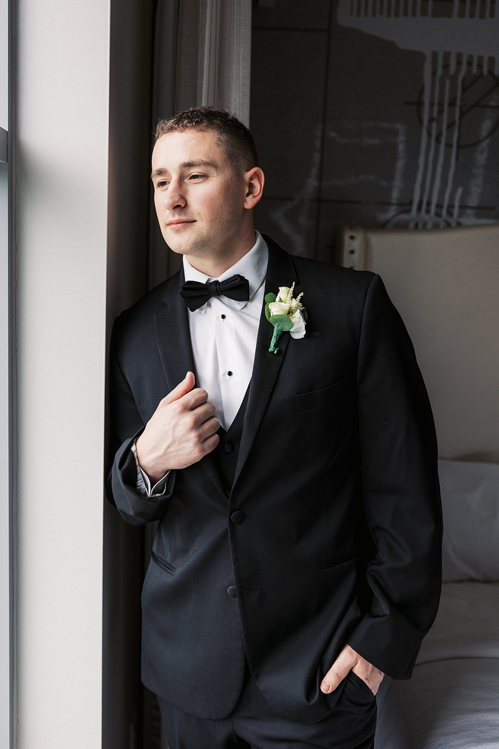 A groom stands in a window holding the lapel of his black suit at a Stonehouse At Stirling Ridge Wedding