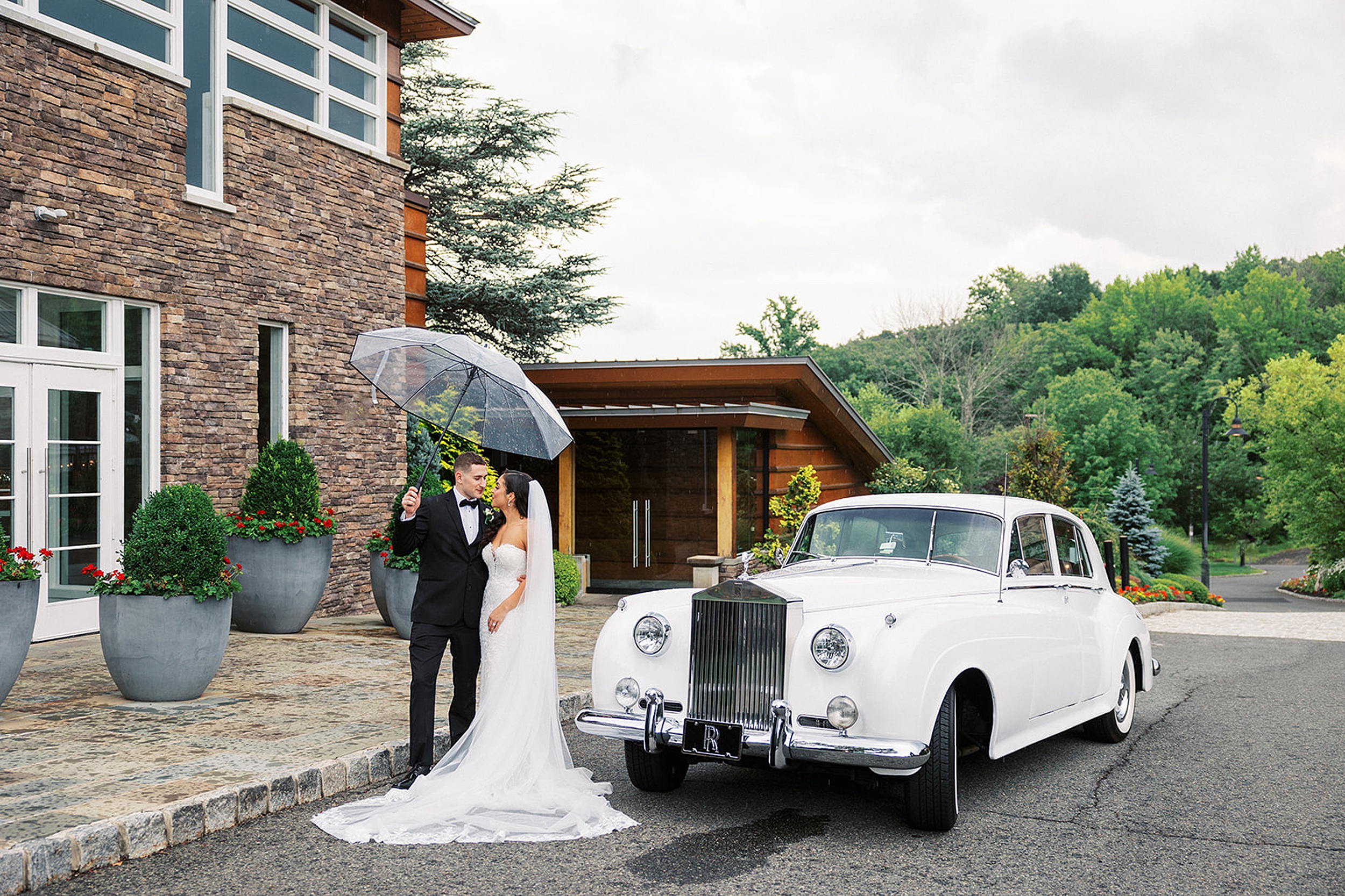 Newlyweds kiss under and umbrella while standing next to a white Rolls Royce limo