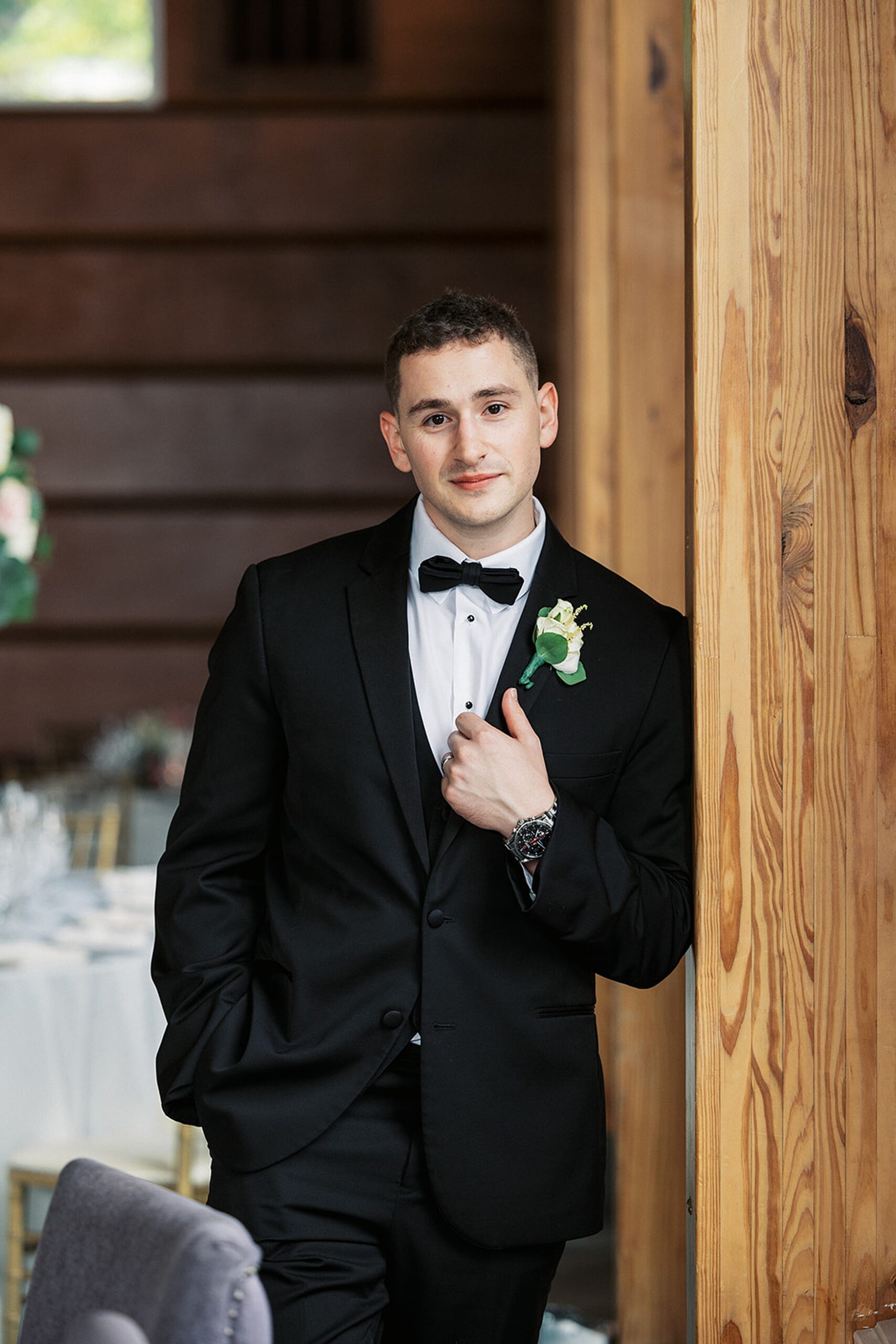 A groom leans against a wooden beam in a wedding reception venue wearing a black suit
