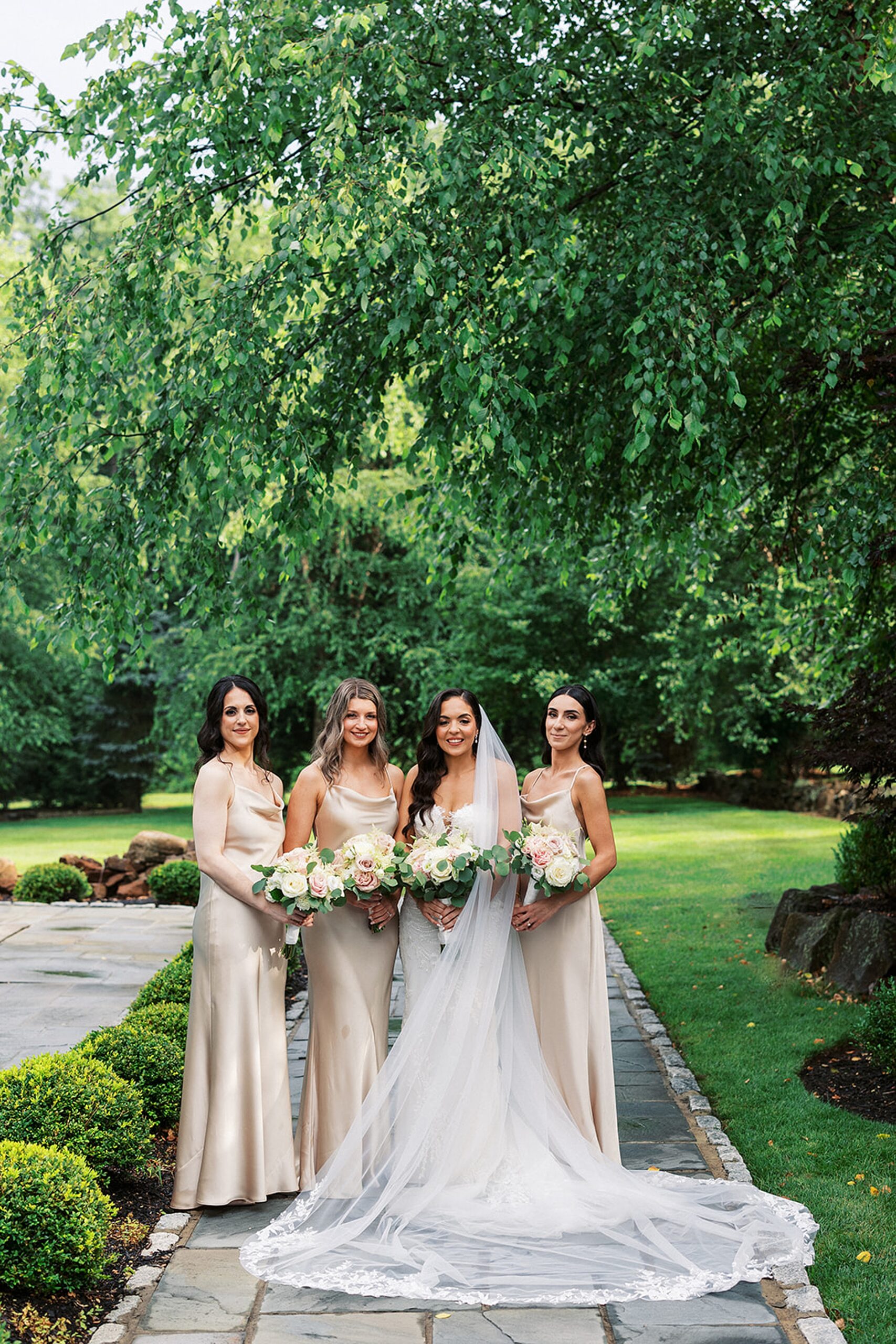 A bride stands in a garden path with her three bridesmaids in gold dresses