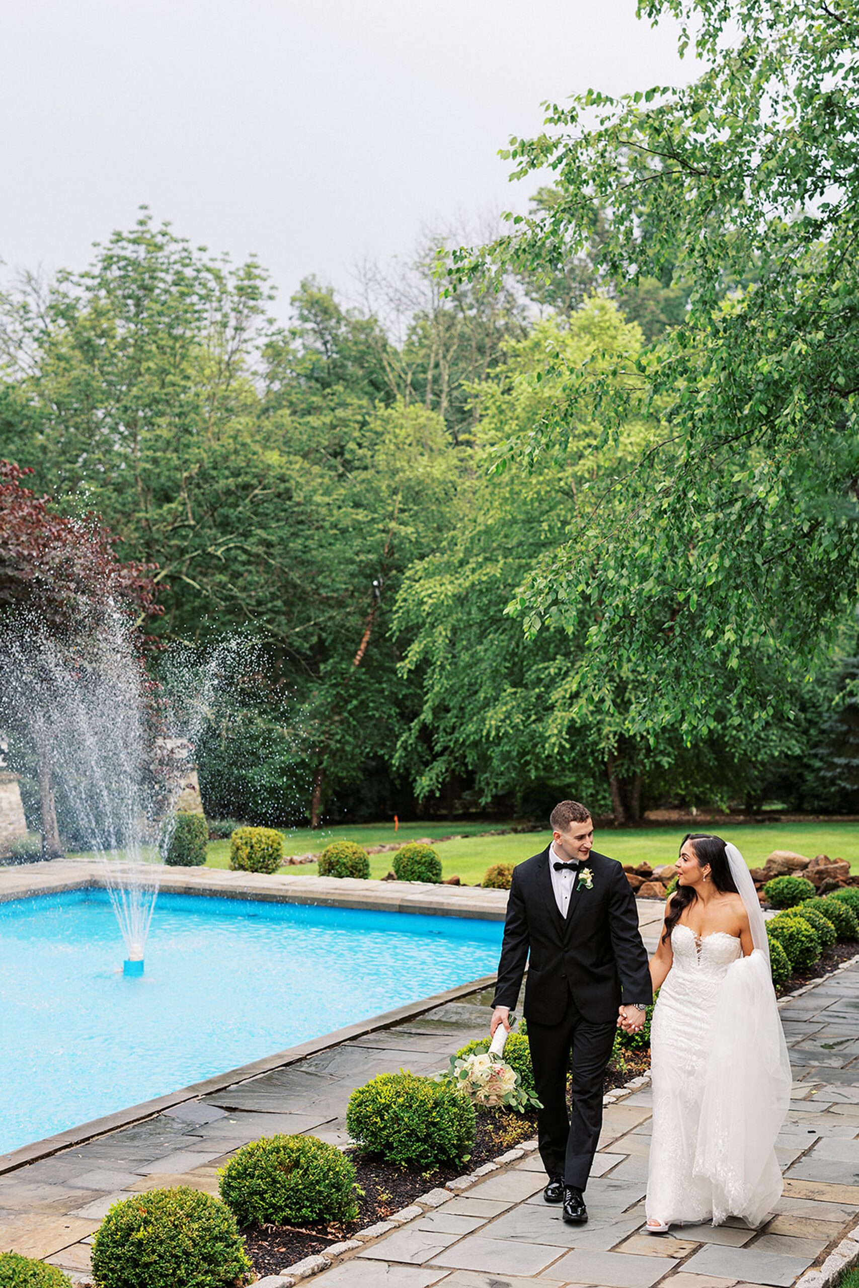 Newlyweds walk past a garden fountain on a stone walkway while holding hands