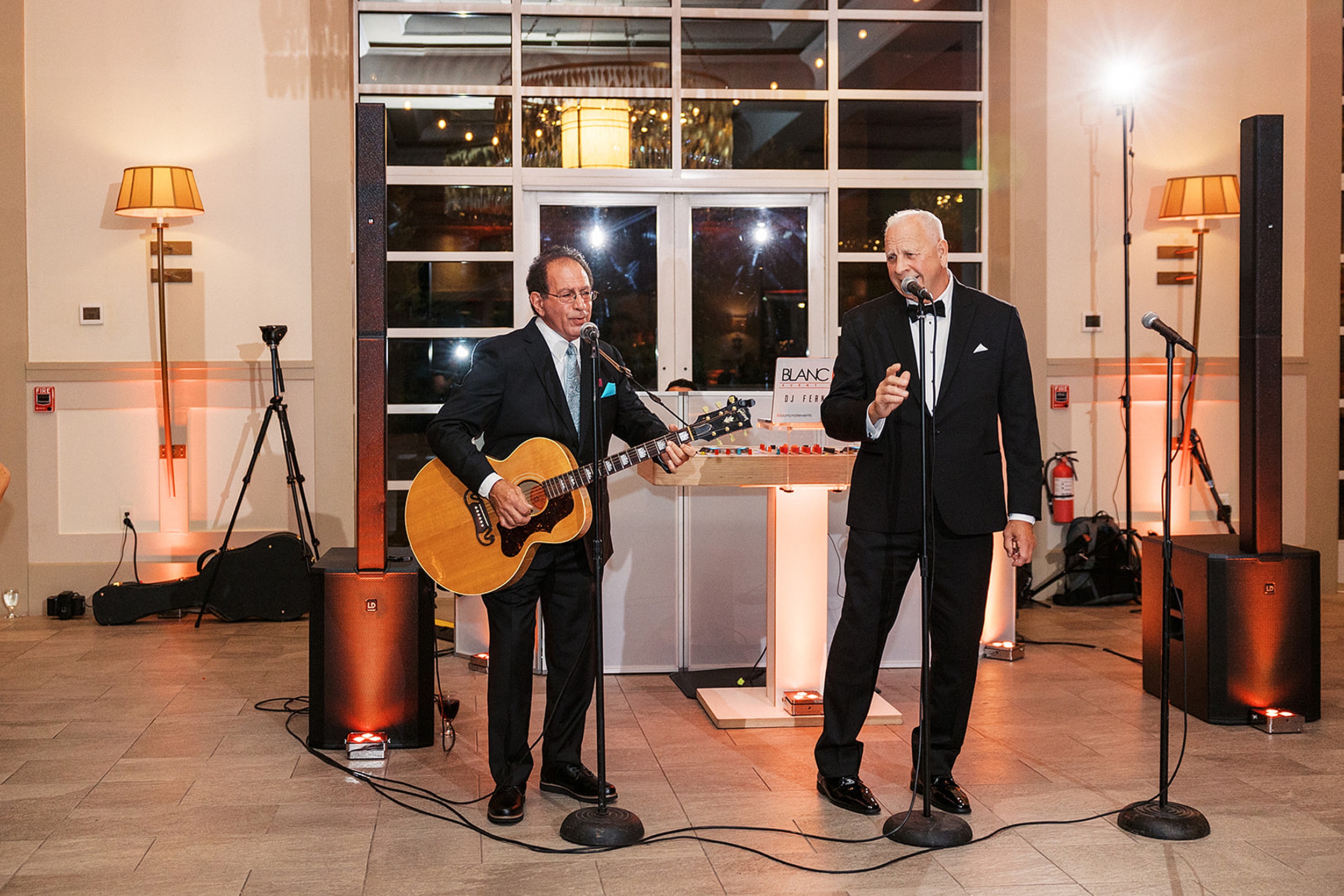 Two men in black suits sing and play guitar at a Stonehouse At Stirling Ridge Wedding