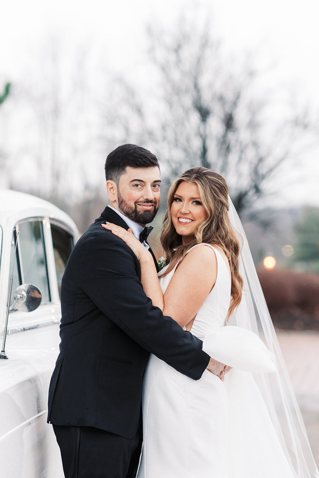 Newlyweds hug while leaning on a classic white car at a Barrow Mansion Wedding