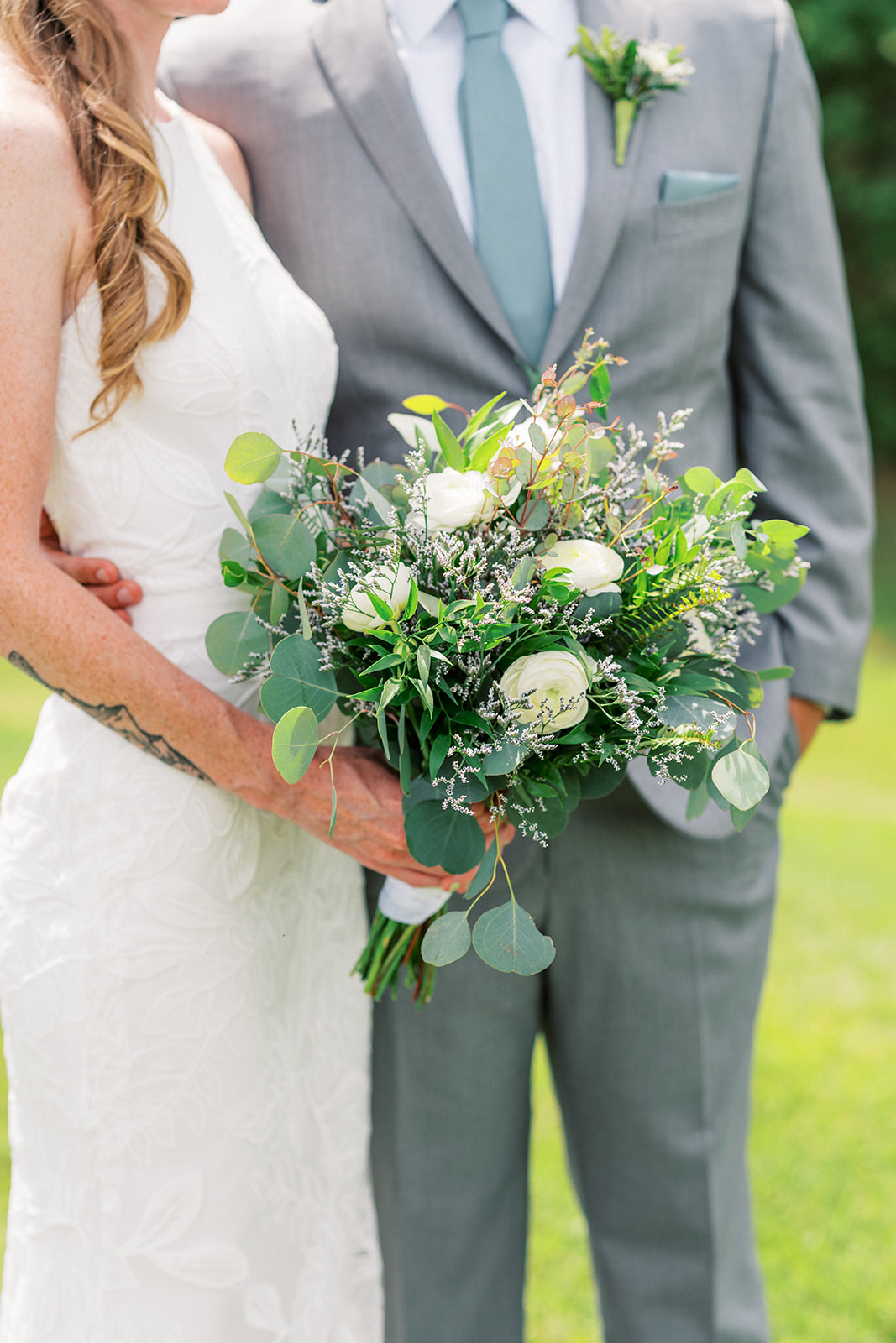 Details of a bride holding her bouquet while tanding in a field with her groom in a grey suit