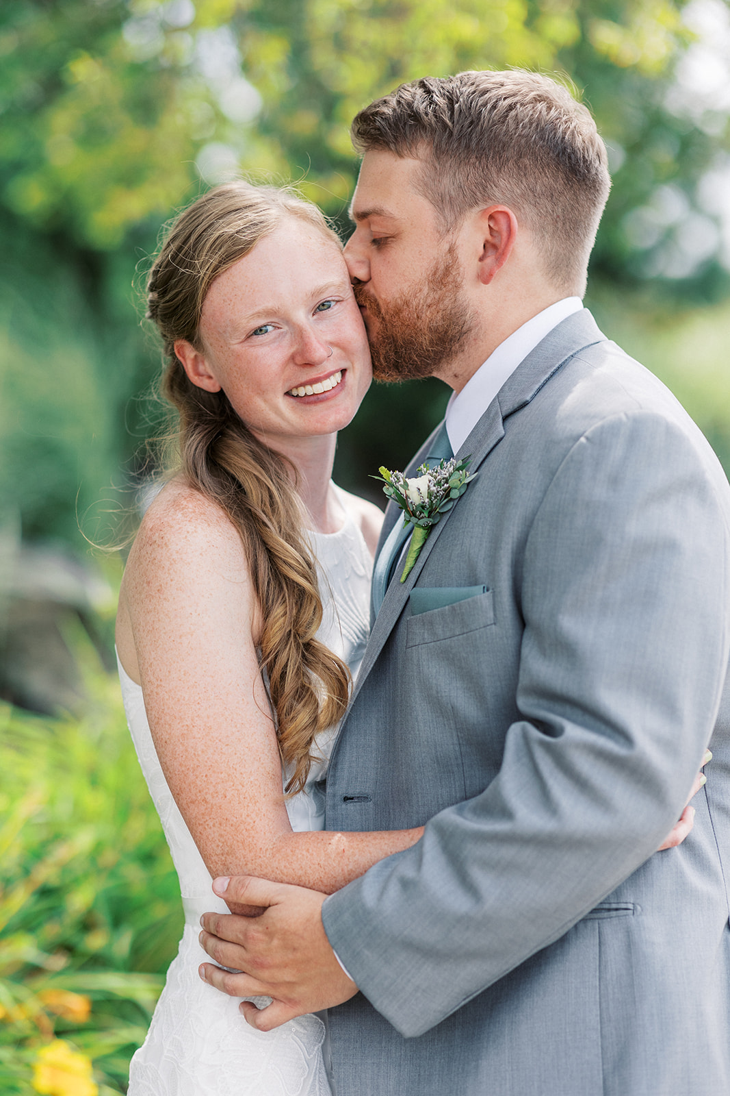 A groom in a grey suit kisses the cheek of his bride in a white lace dress at a Galloping Hill Wedding