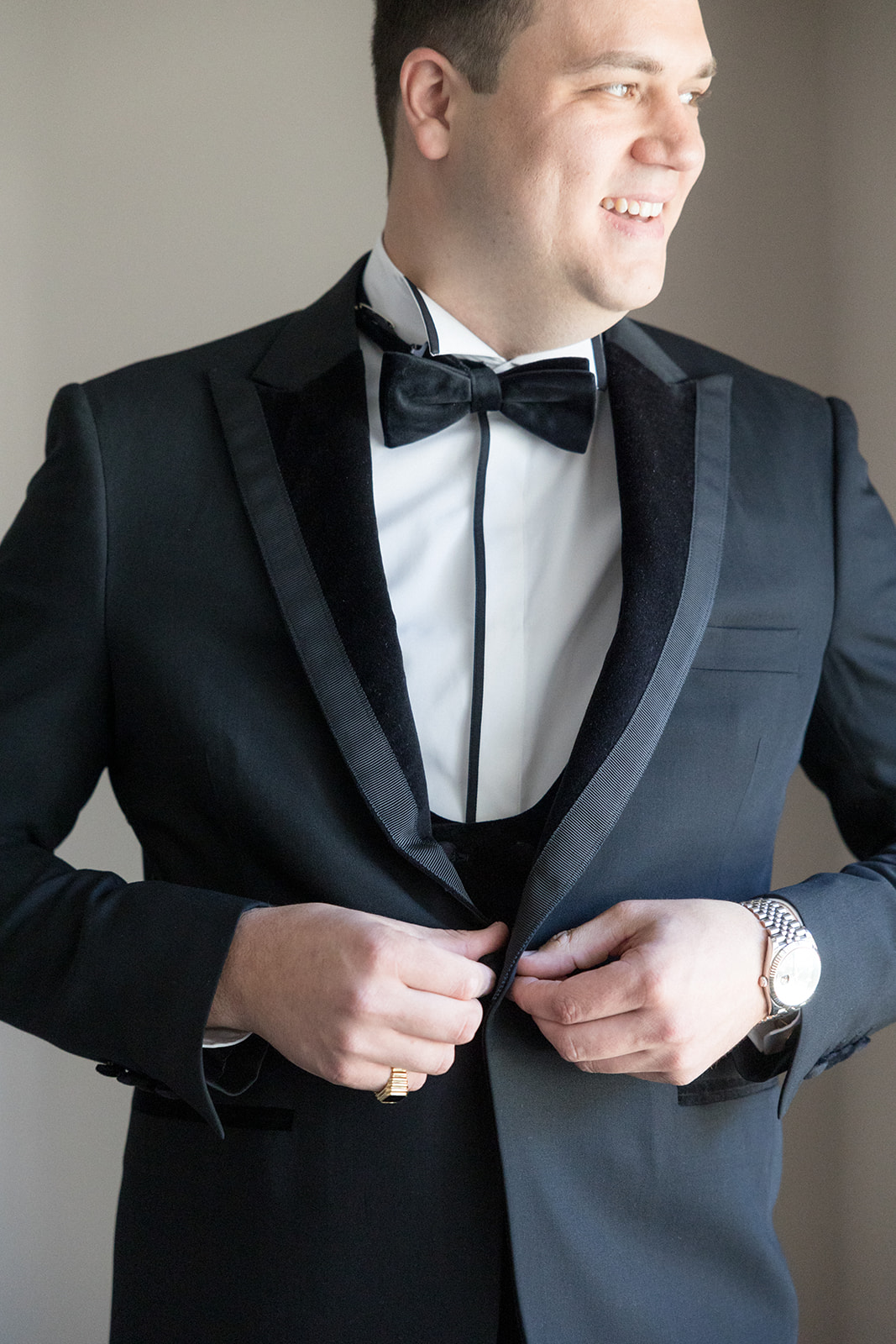 A groom adjusts his buttons on his tuxedo jacket while standing and looking out a window at a James Ward Mansion Wedding