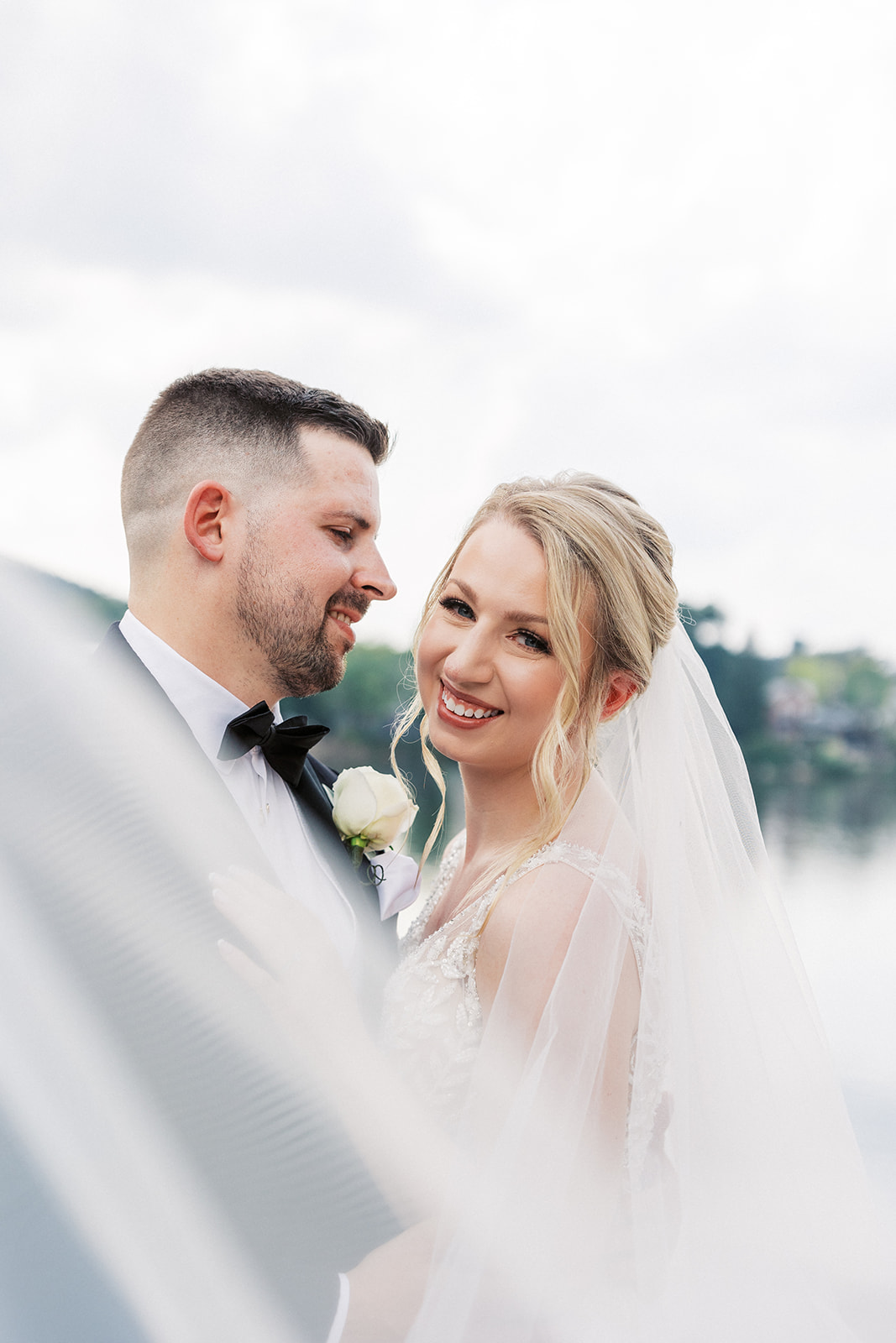 Newlyweds laugh while standing in a waterfront park as the veil blows in the wind around them