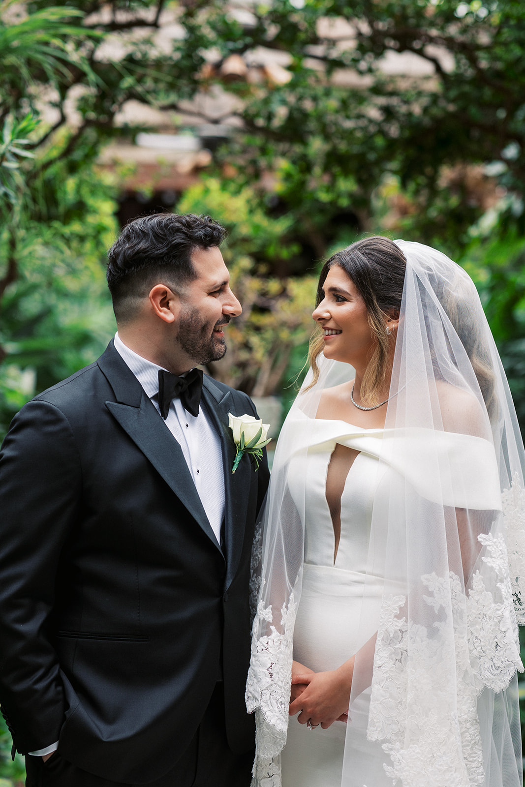 Newlyweds stand in a garden while wearing a black tuxedo and white silk dress with long embroidered veil at The Royal Manor Wedding venue
